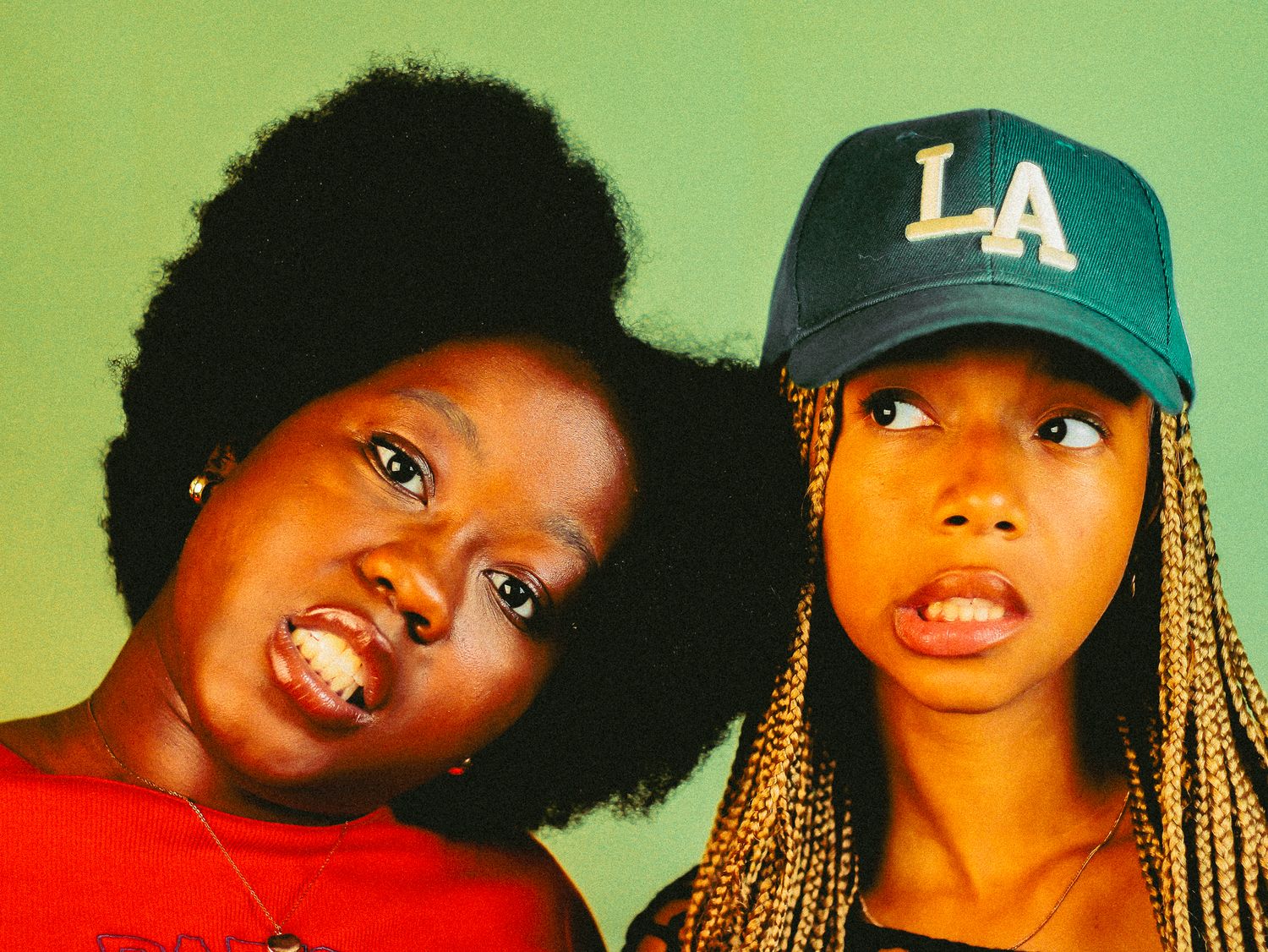 Two young Black women making funny faces together, one with natural afro in red top, other in LA cap.