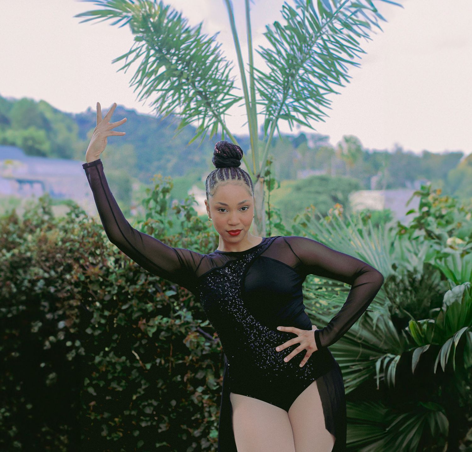A performer in a sparkly black dance costume poses with raised arms among lush greenery.