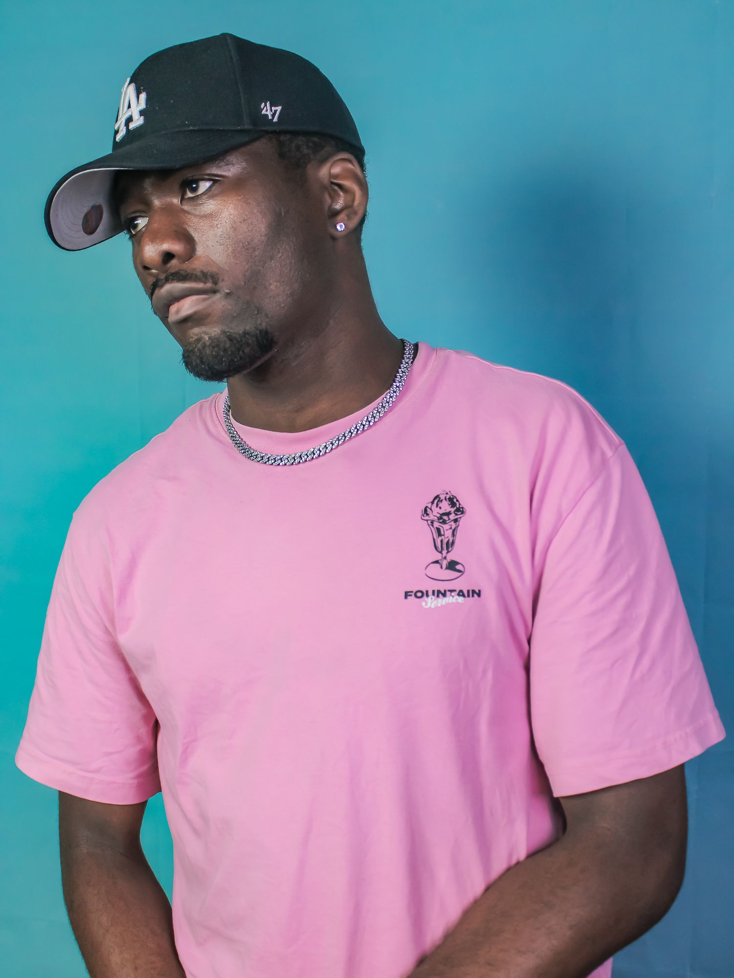 Stylish man in pink Fountain graphic tee and black LA cap looks away against teal backdrop.