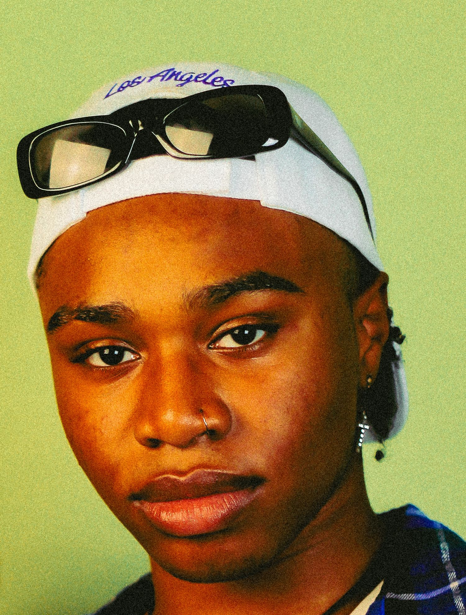 Close-up portrait of young person with white Los Angeles cap and sunglasses on forehead, blue plaid shirt.