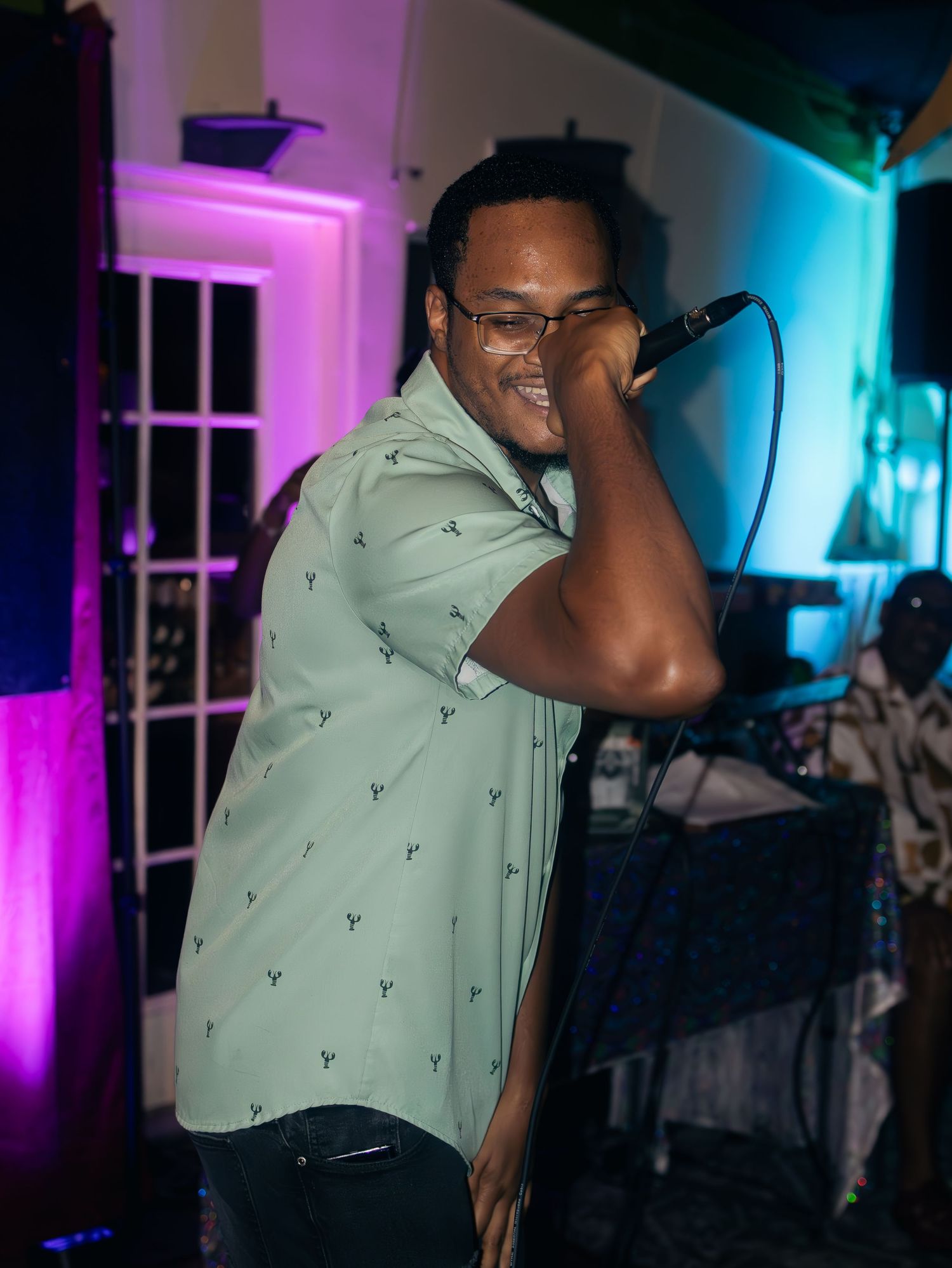 Performer in a light green button-up shirt singing passionately on stage with colorful lighting effects.