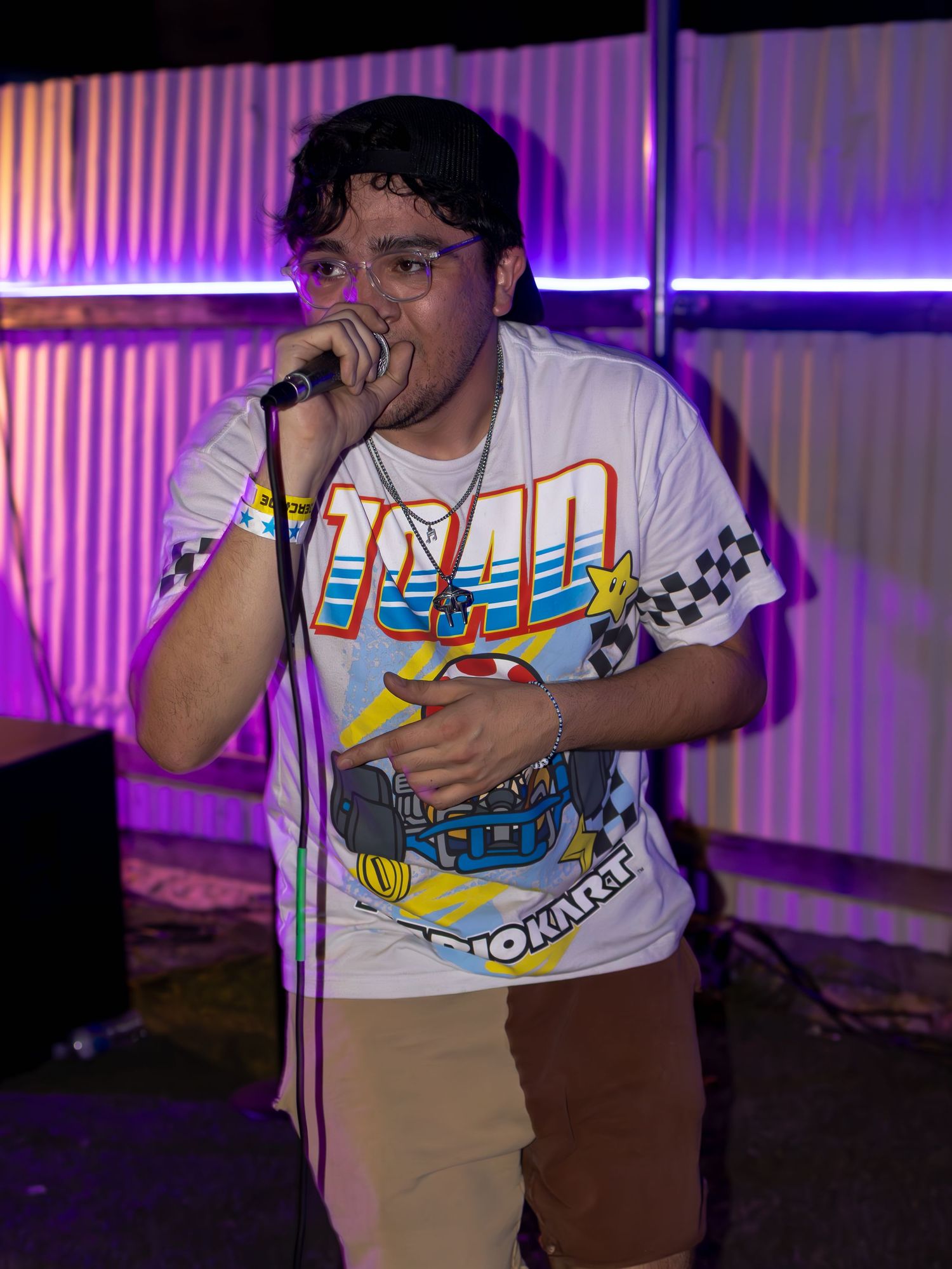 Concert performer wearing gaming t-shirt and baseball cap performing under vibrant purple stage lights.