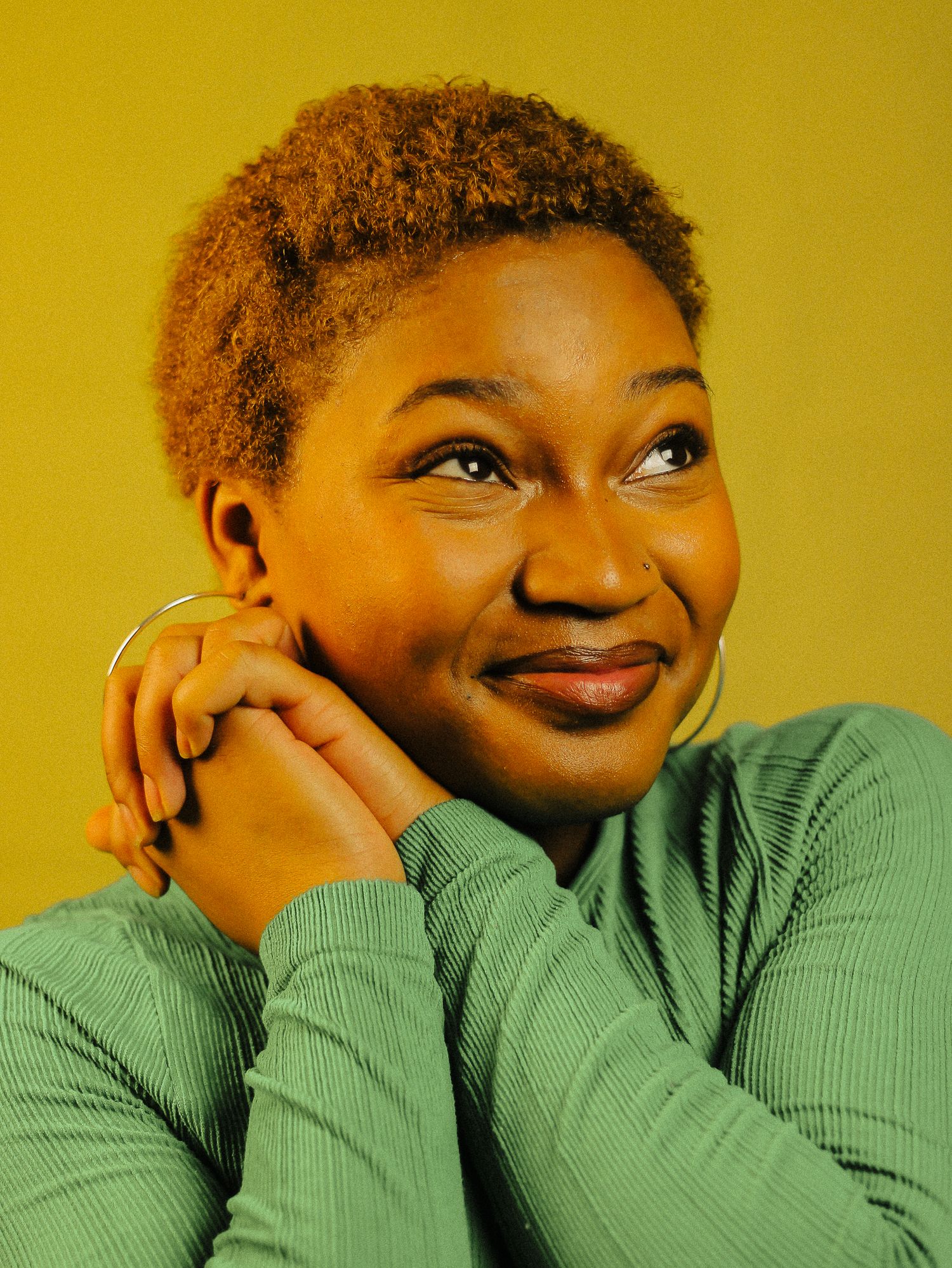 A young Black woman in a green ribbed turtleneck smiles thoughtfully against a yellow background.