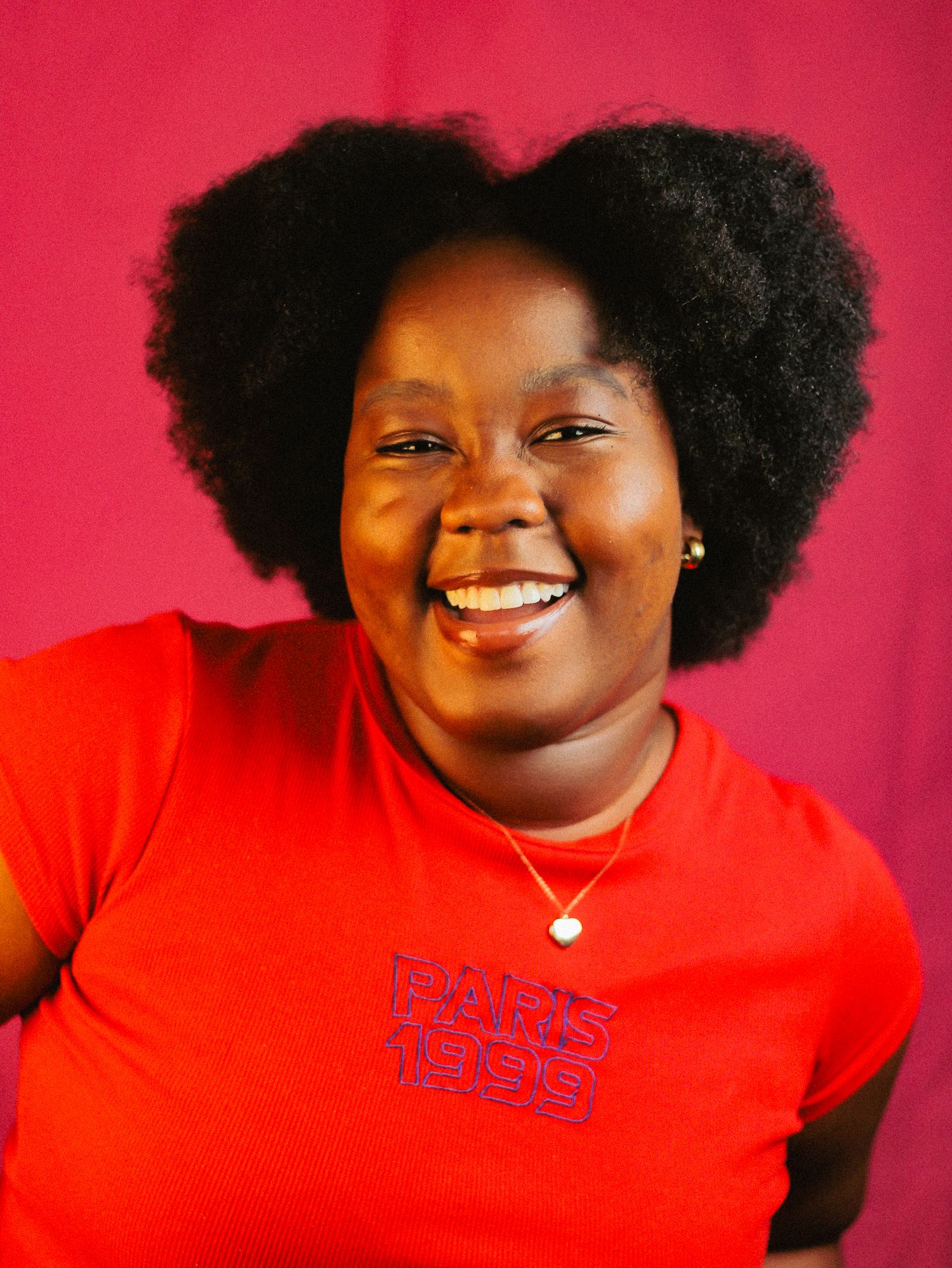 Smiling Black woman with natural afro hair wearing a red Paris 1999 shirt against a deep pink background.