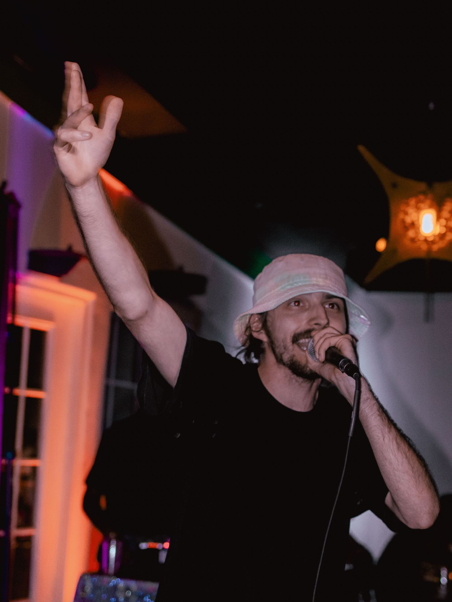 Performer in white cap gesturing dramatically during hip hop performance on dimly lit stage.