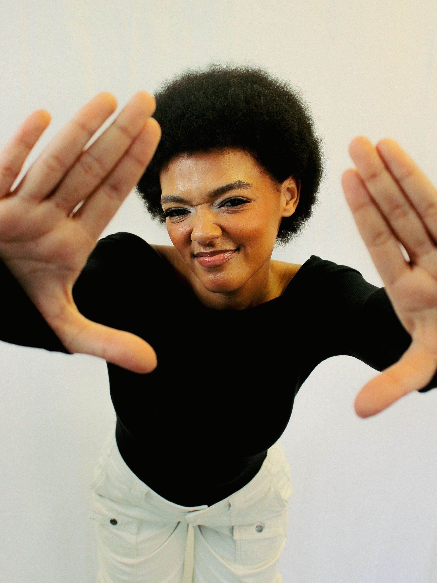 Person in black top and white pants making dynamic hand gestures against light background.