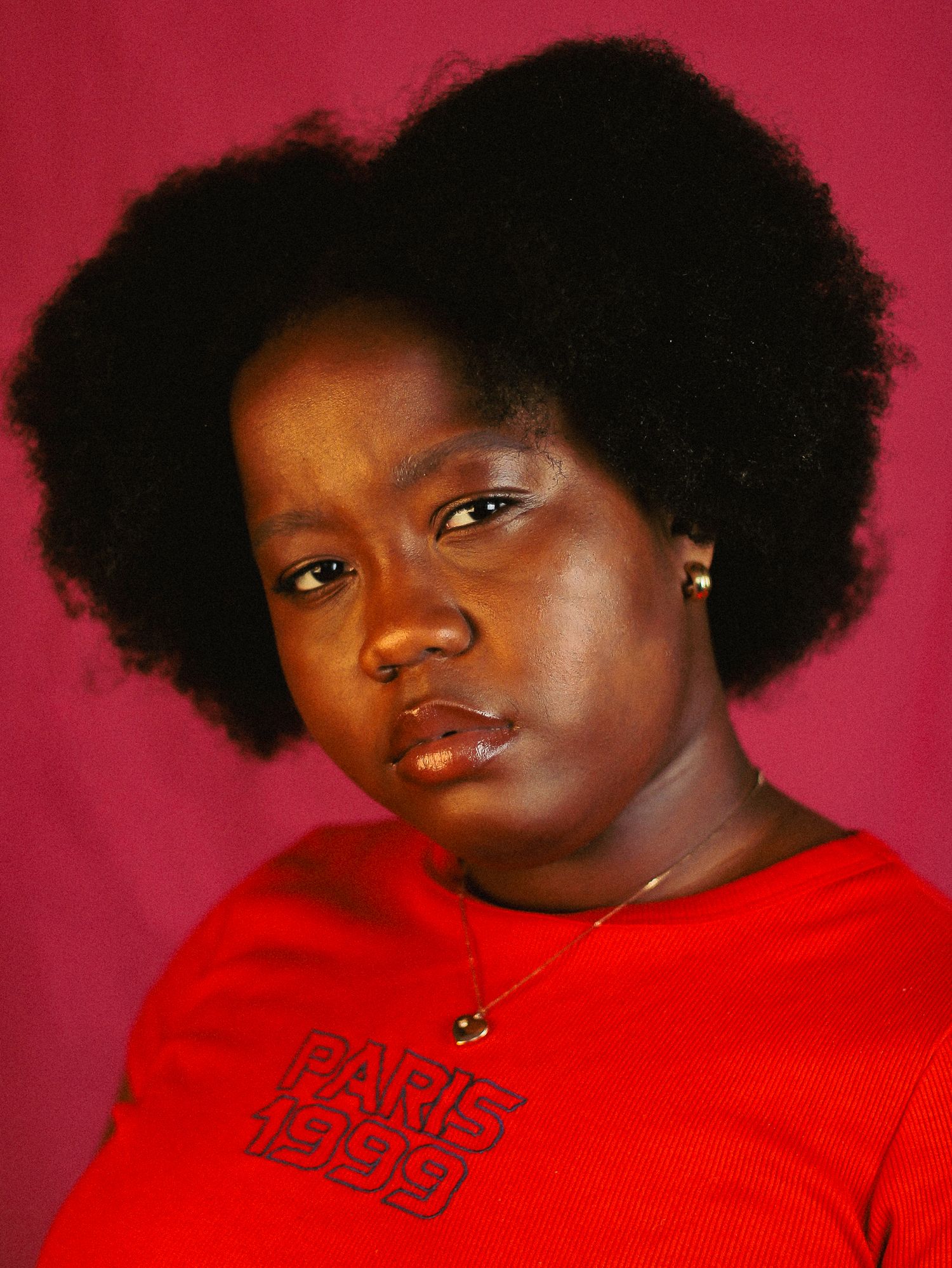 Black woman with voluminous natural afro hair in a red Paris 1999 shirt with a serious expression, pink background.