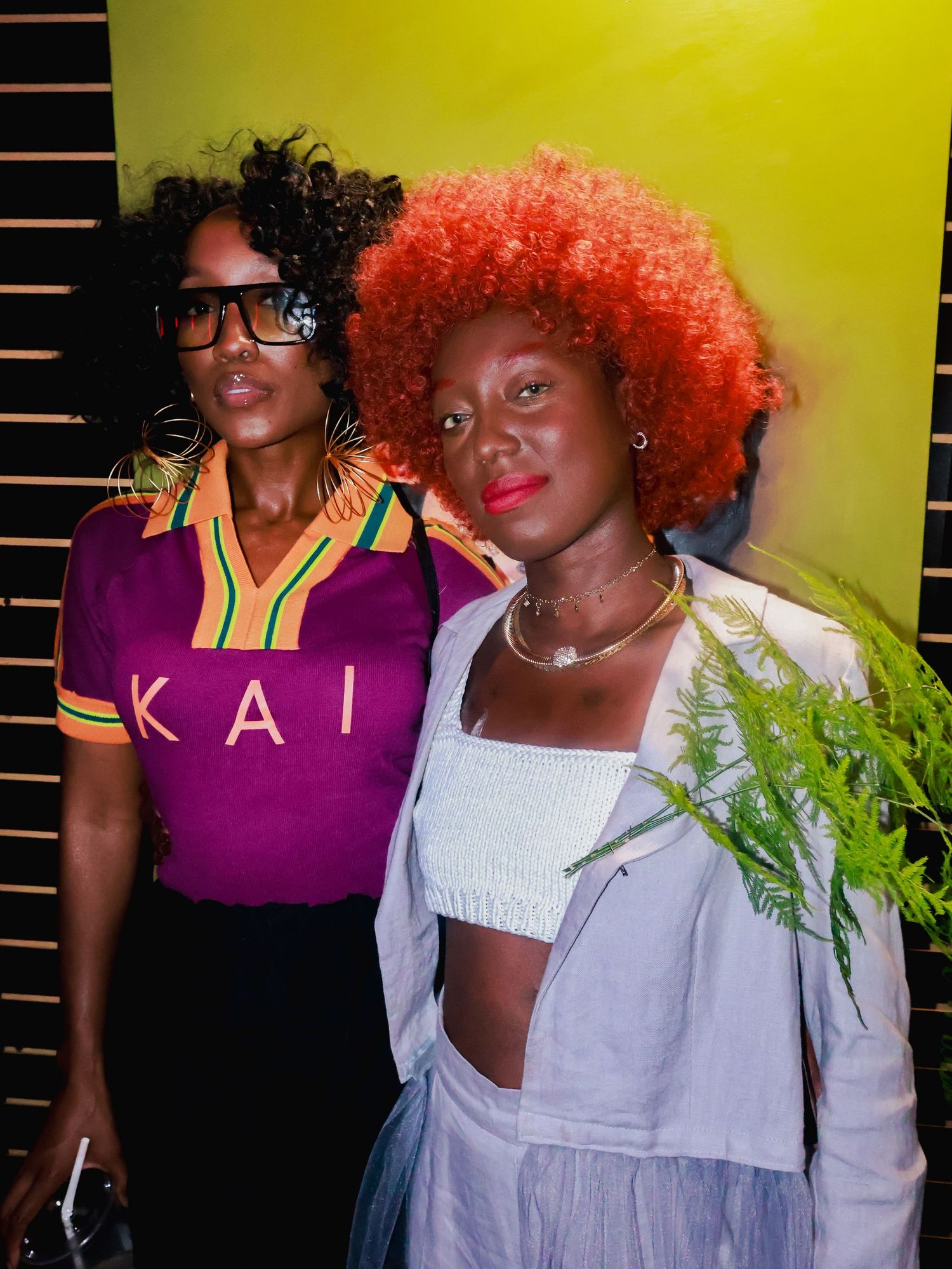 Two people smile and pose together in casual outfits against a bright yellow background.
