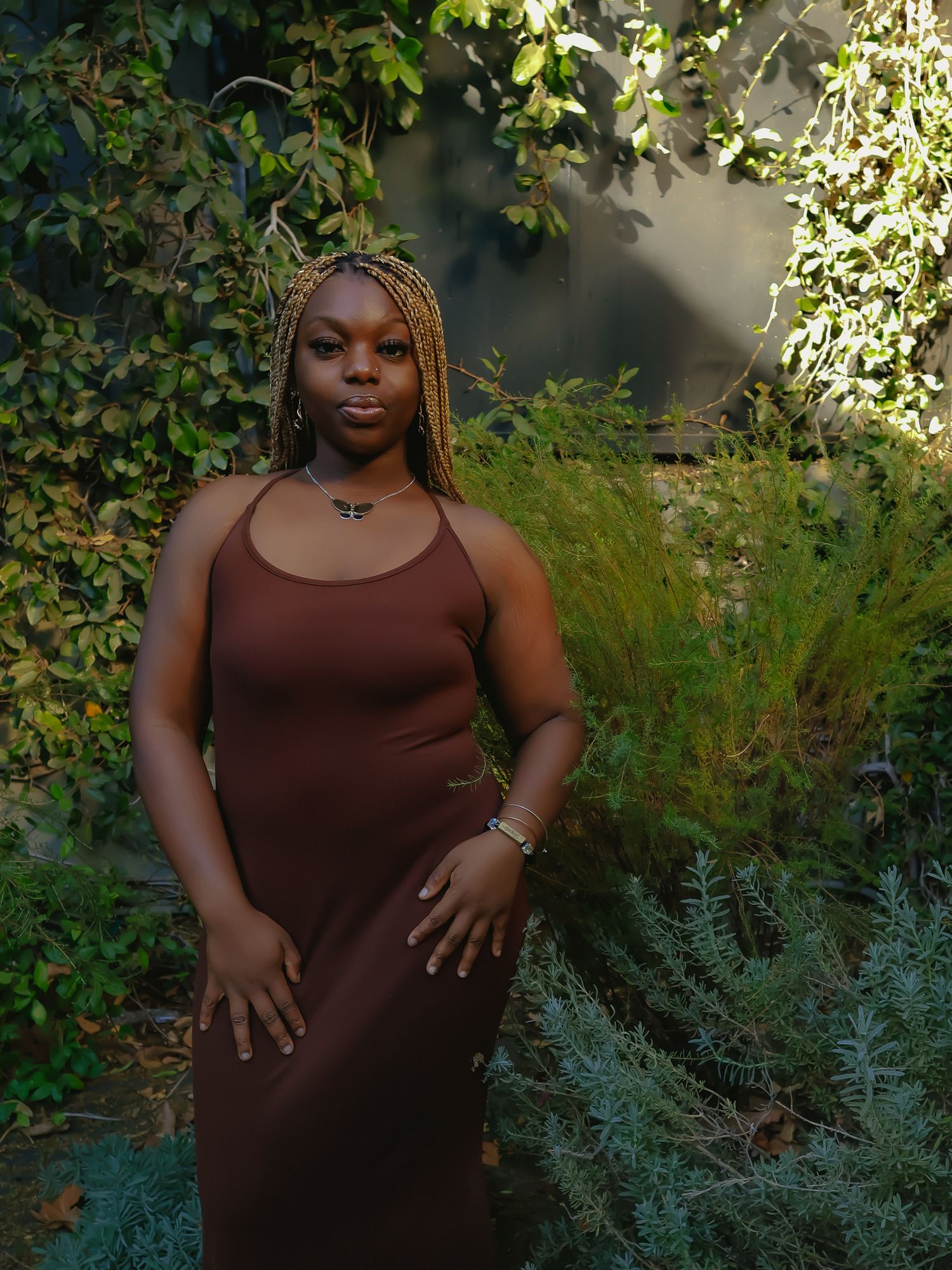 Figure in brown sleeveless top standing in garden setting with decorative plants.
