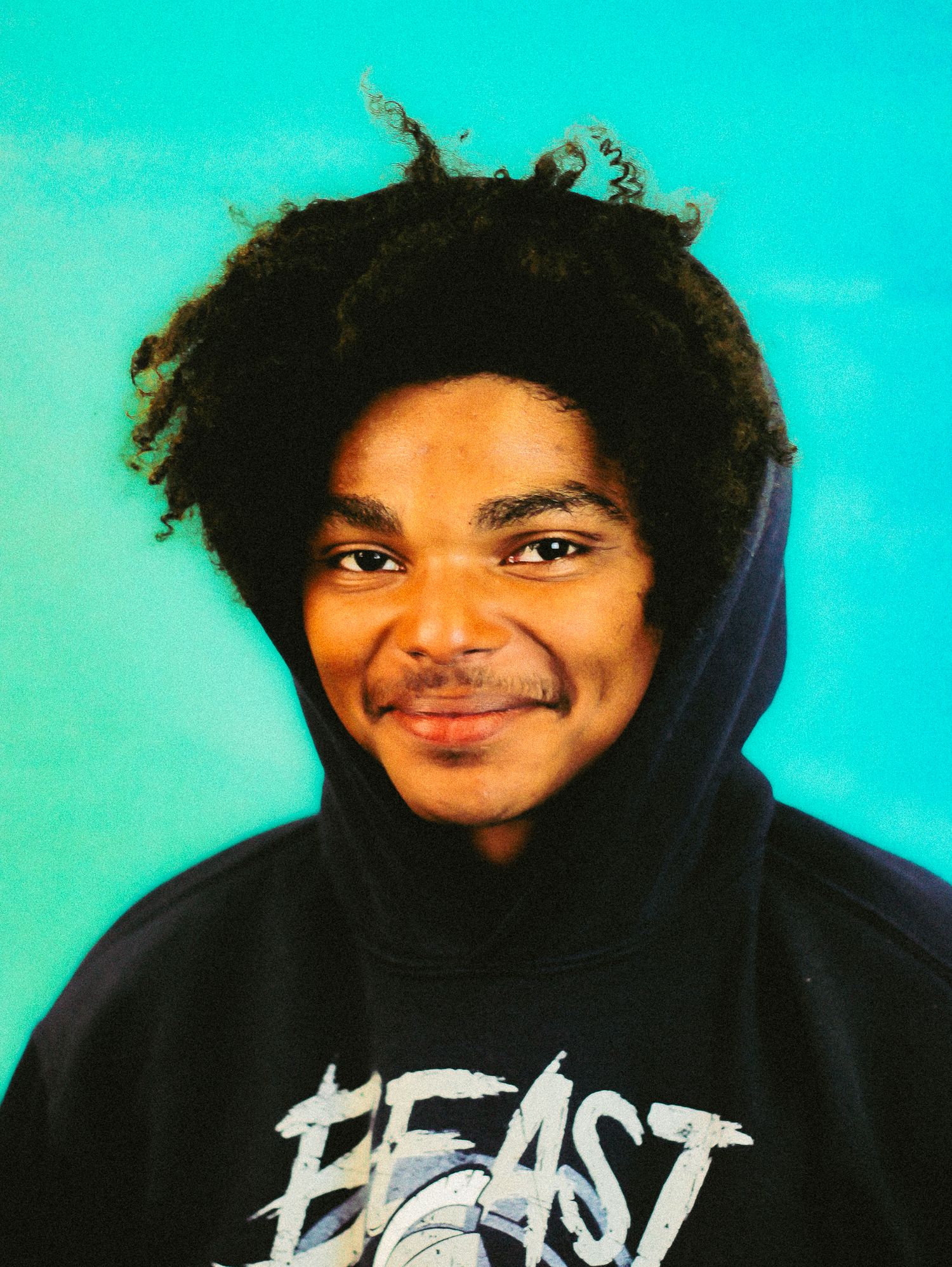 Smiling young man with curly afro wearing black Beast hoodie against bright turquoise background.