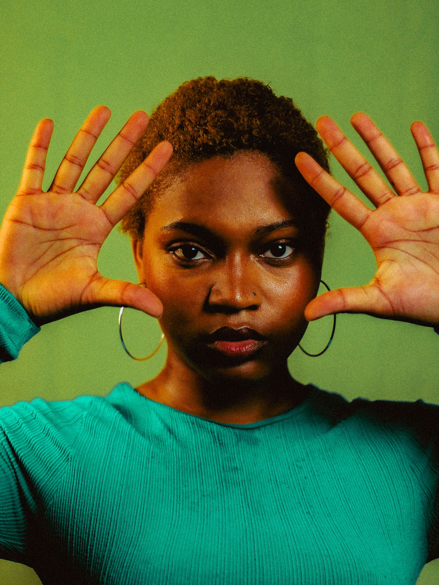 A young Black woman in a teal sweater holds both hands up facing forward against a green background.