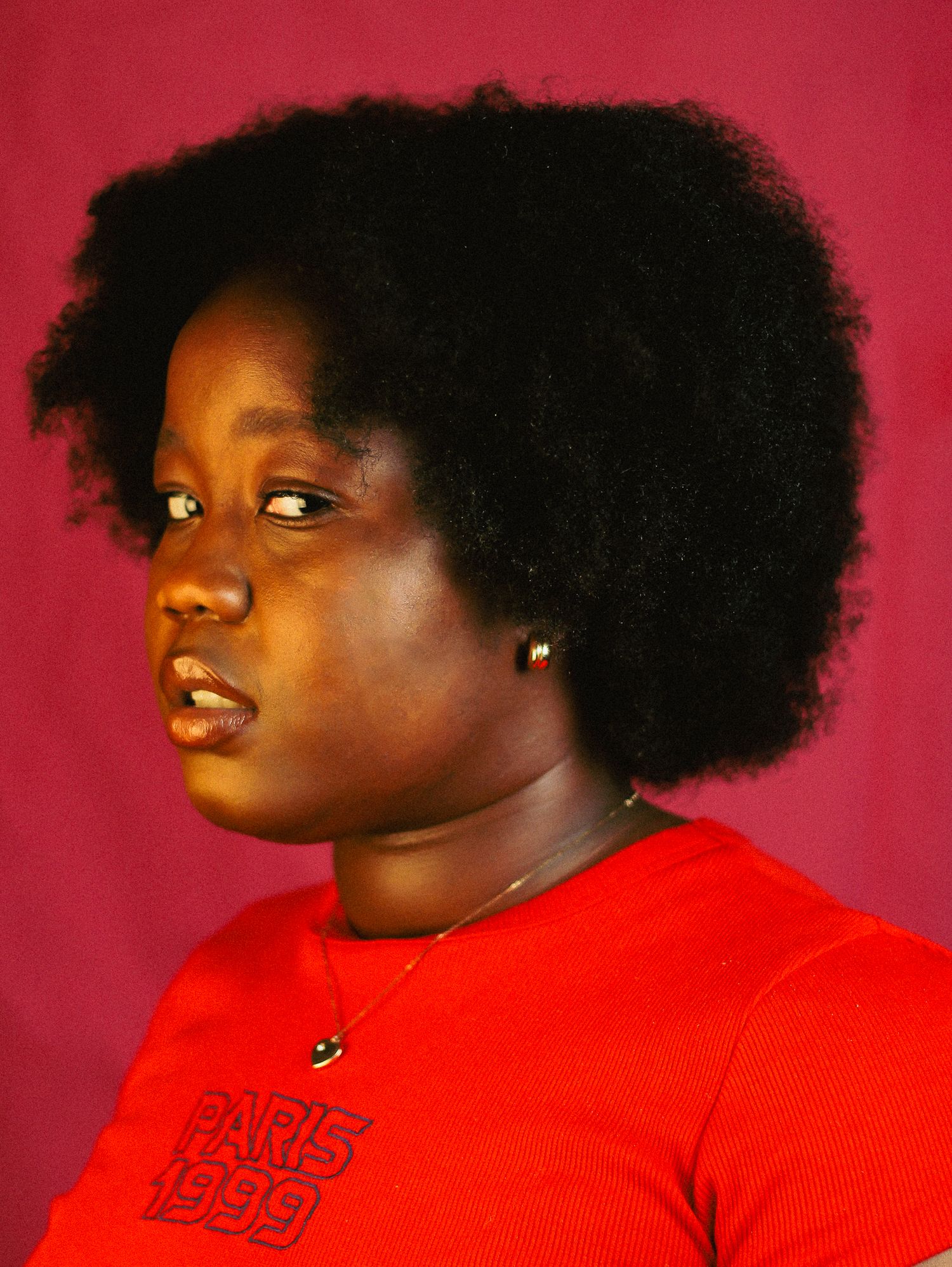 Black woman with natural afro hair in a red Paris 1999 t-shirt gazing to the side against a pink backdrop.