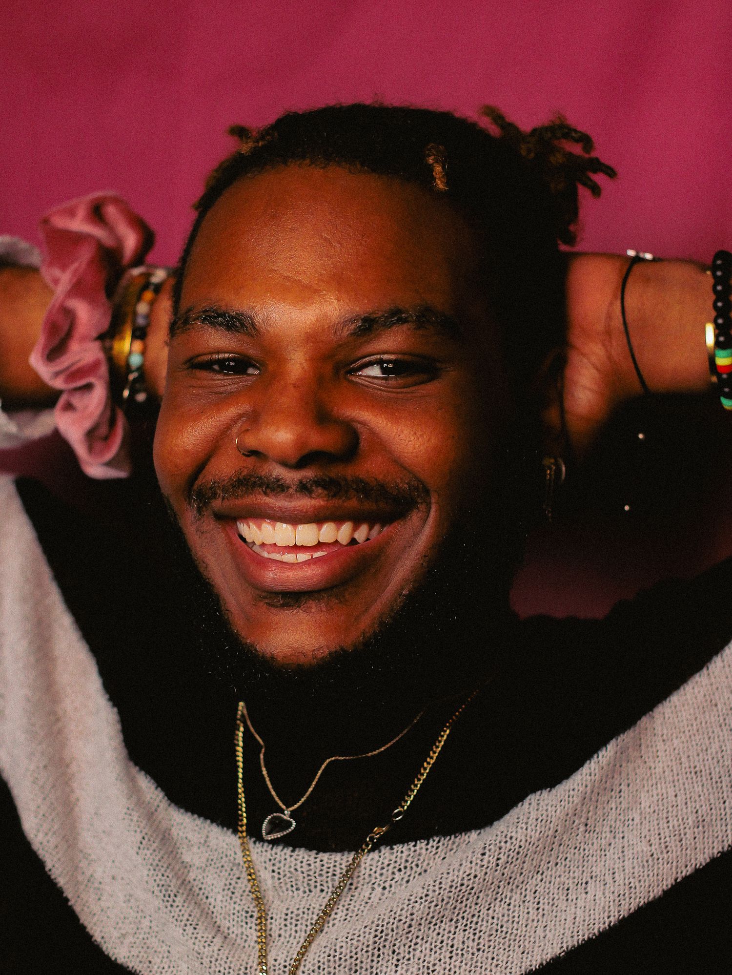 Portrait against red background showing joyful expression and gray sweater with gold necklace.