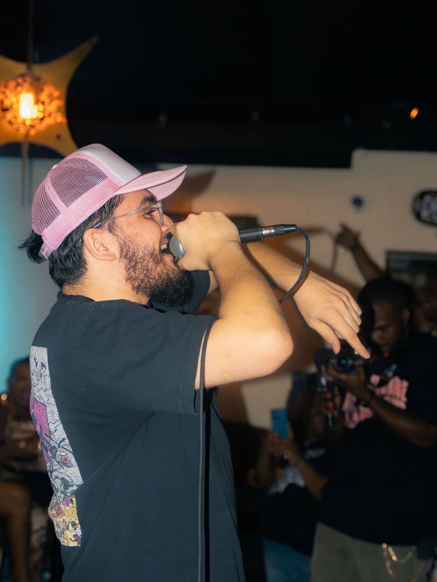 Performer in pink cap and black t-shirt energetically singing into microphone on stage with colorful lighting.