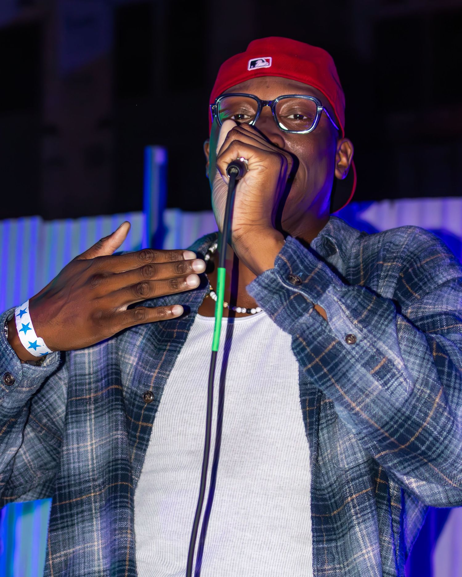 Artist in plaid shirt and glasses performs with microphone under blue stage lighting.