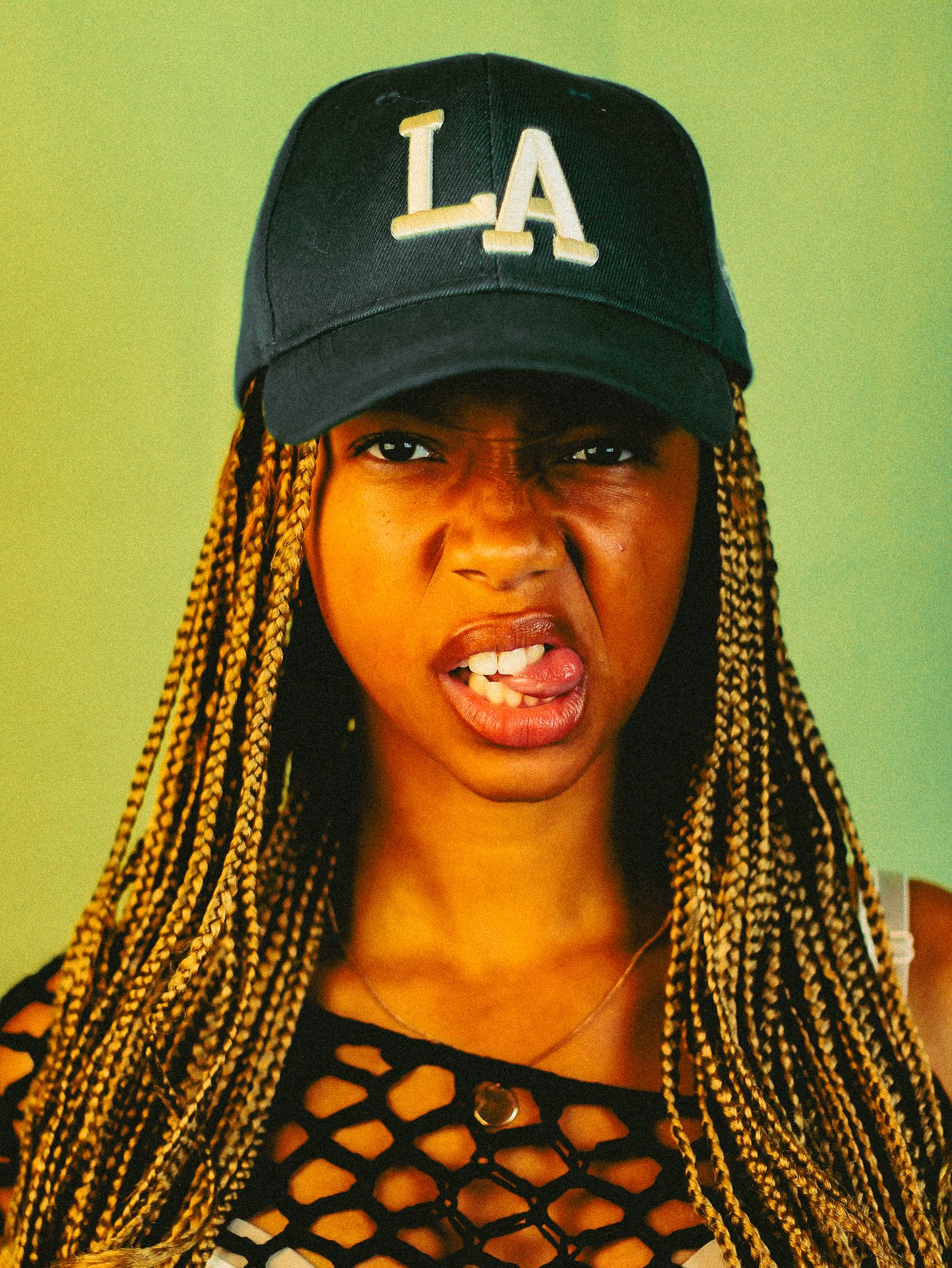 Young woman wearing an LA baseball cap and mesh top makes bold expression against green background.
