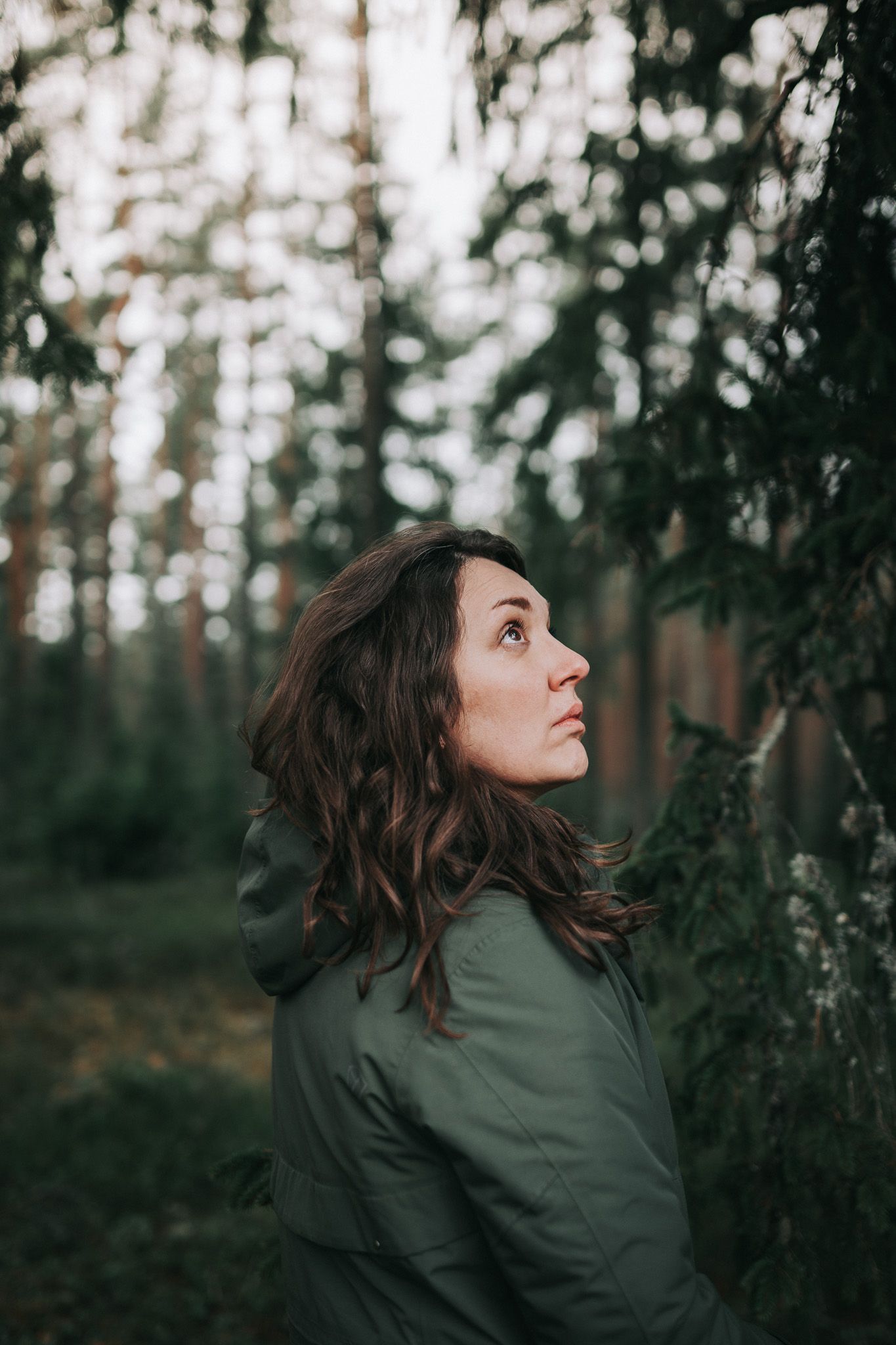 Person in dark green jacket looks up thoughtfully in moody forest setting at dusk.