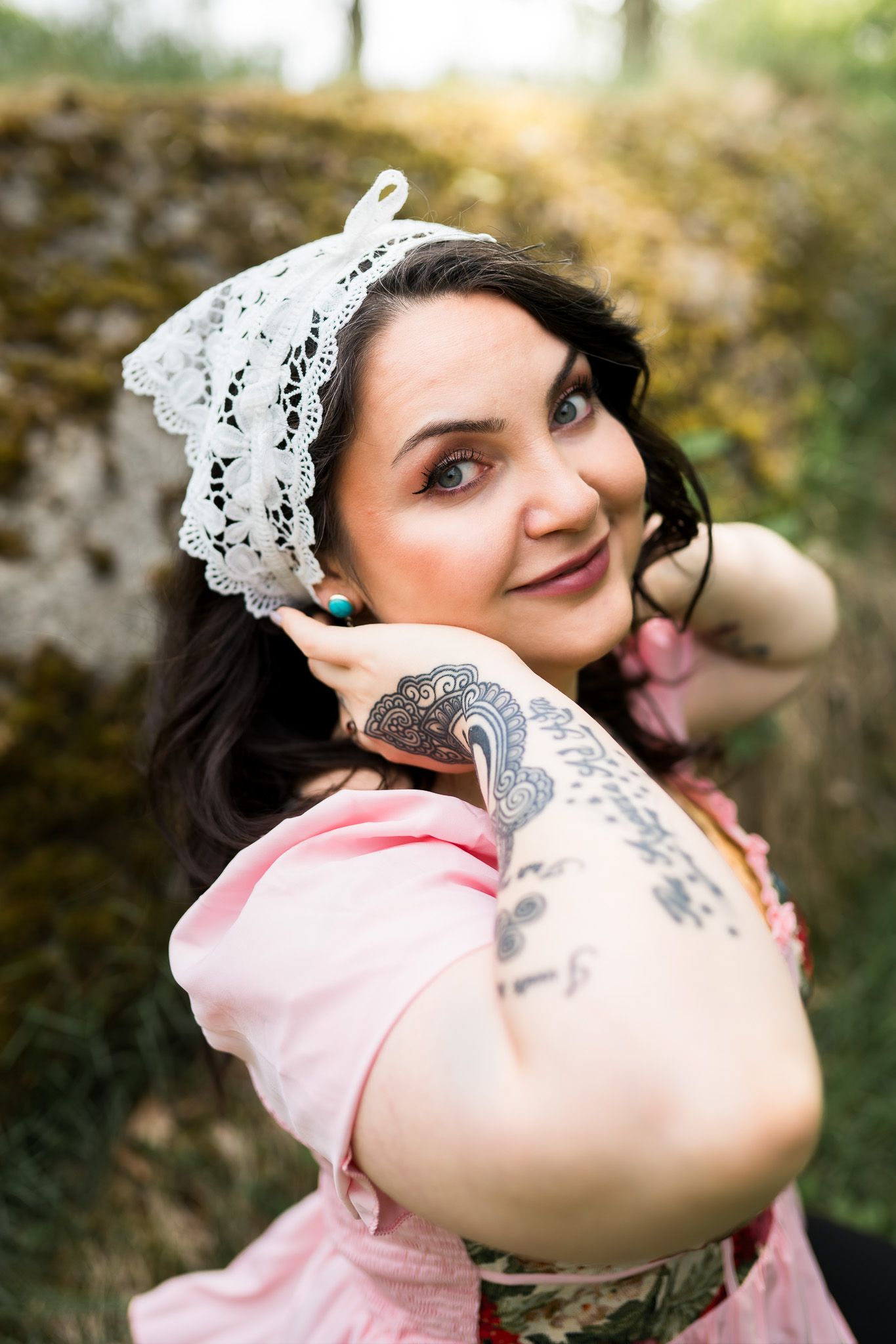 A vintage-inspired portrait featuring a white lace hair accessory, pink dress and intricate arm tattoo in an outdoor setting.