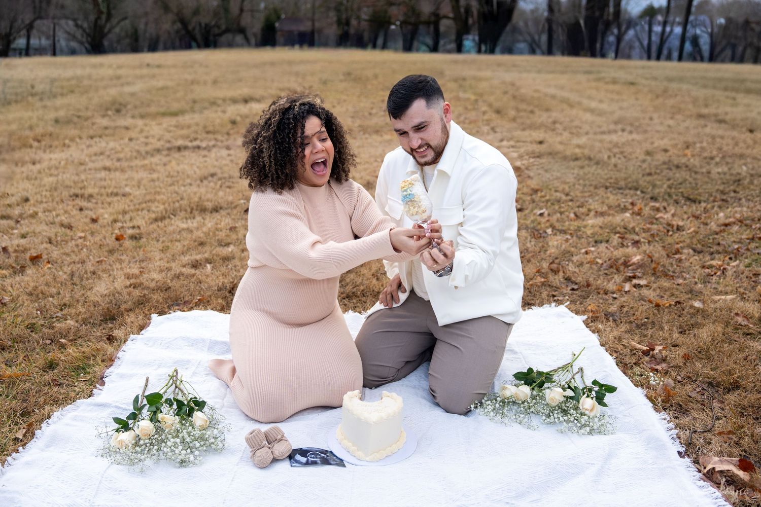 A couple celebrates on a white blanket in a field with floral arrangements during a gender reveal.