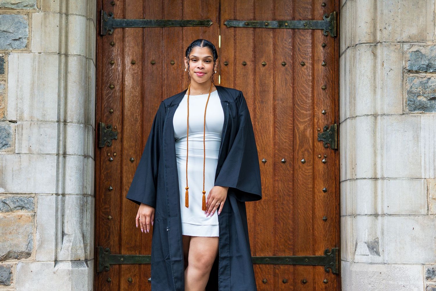 Graduate in black gown and white dress posing in front of ornate wooden door.