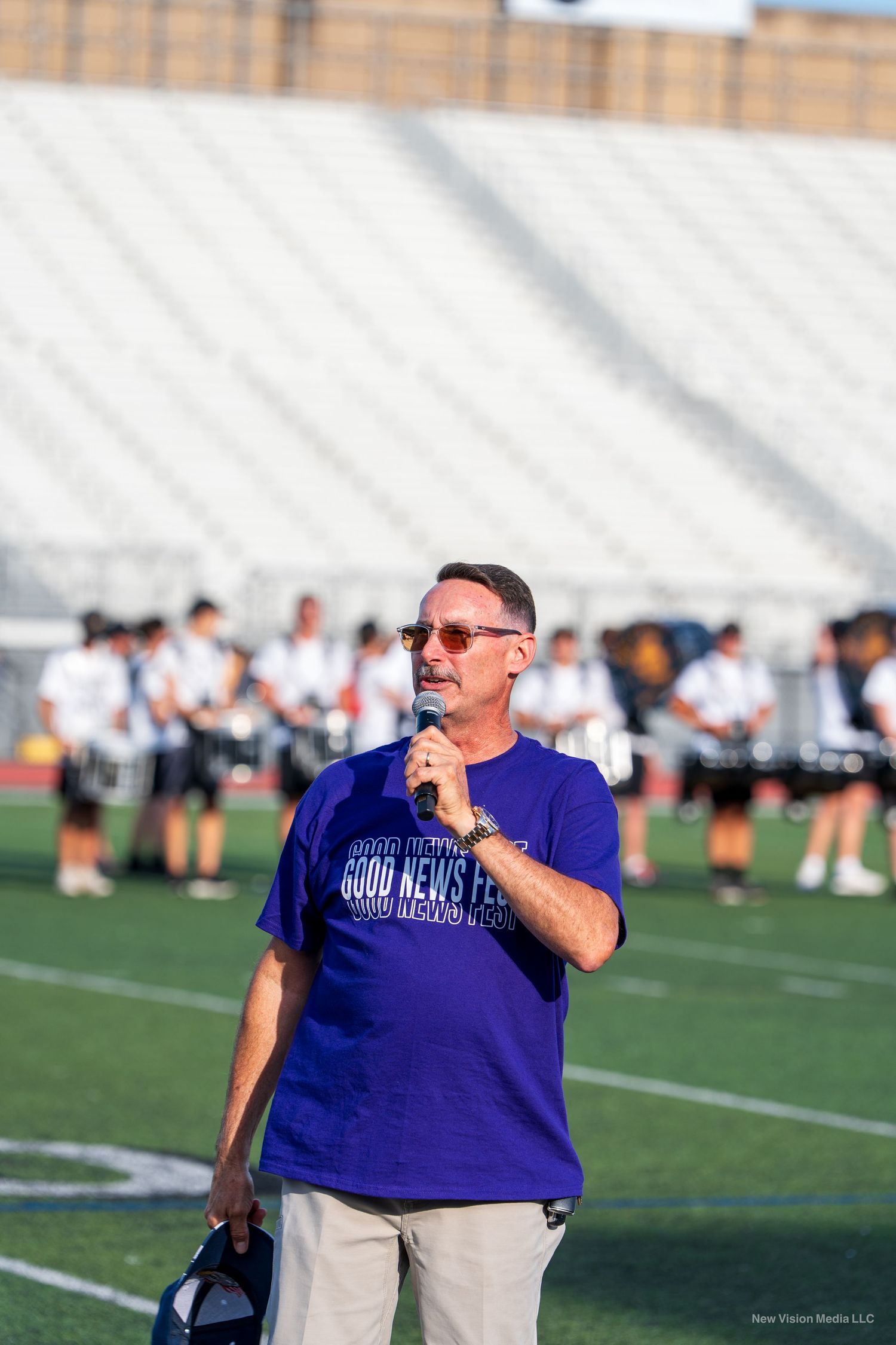Someone in a purple shirt speaks into a microphone on a football field.