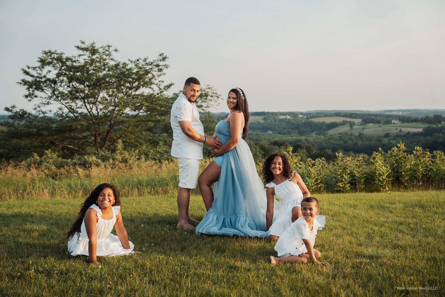 Family portrait in casual white outfits against scenic hilltop sunset view.