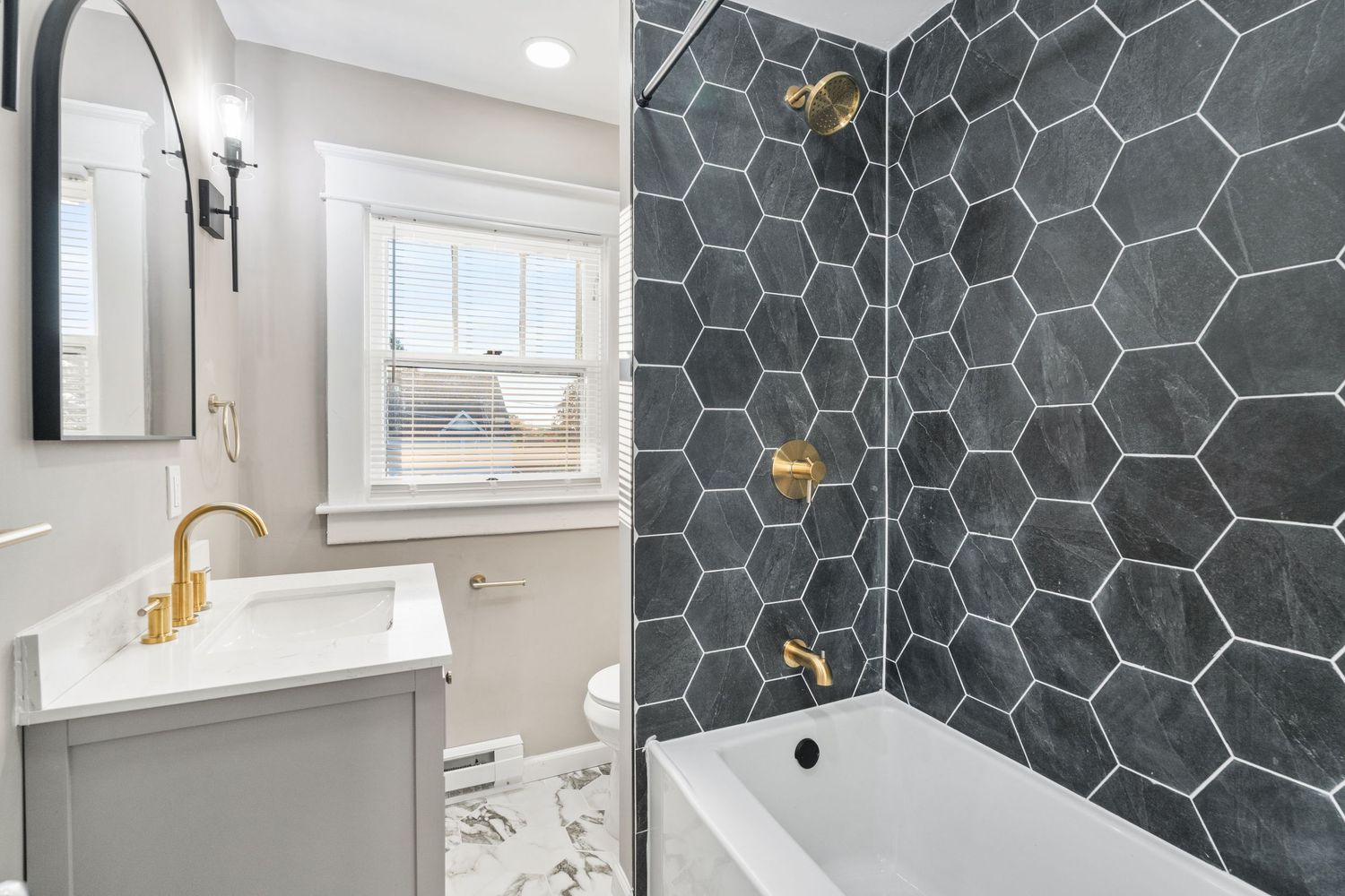 Contemporary bathroom with hexagonal grey tile, gold fixtures, and a white vanity beside a bathtub.
