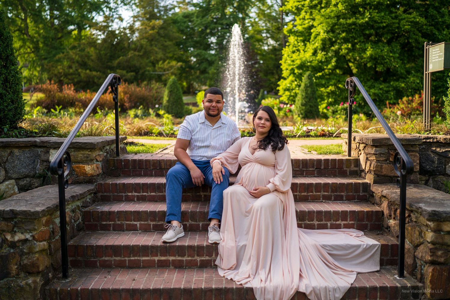 A couple sits together on stone garden steps with a fountain and lush greenery in the background.