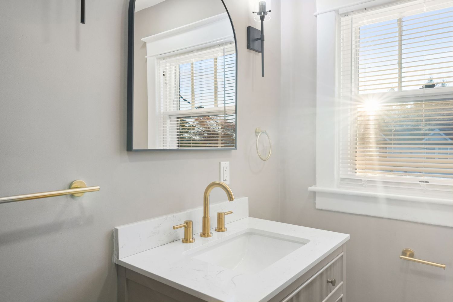 Bright bathroom interior with gold faucet, white vanity, and large framed mirror below window.