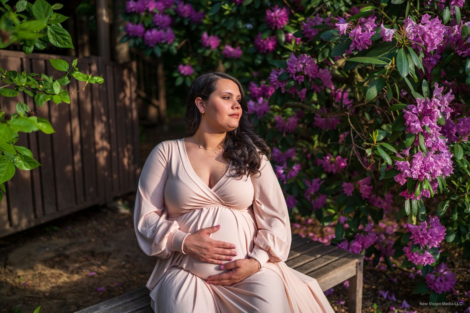Maternity portrait in pink dress against vibrant purple flowers in garden setting.