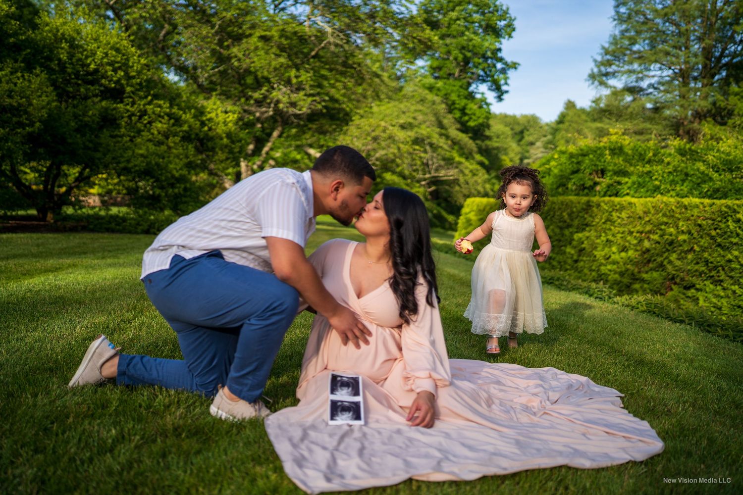 A couple sitting on a blanket in a park looking at a sonogram while a young child in a pink dress stands nearby.