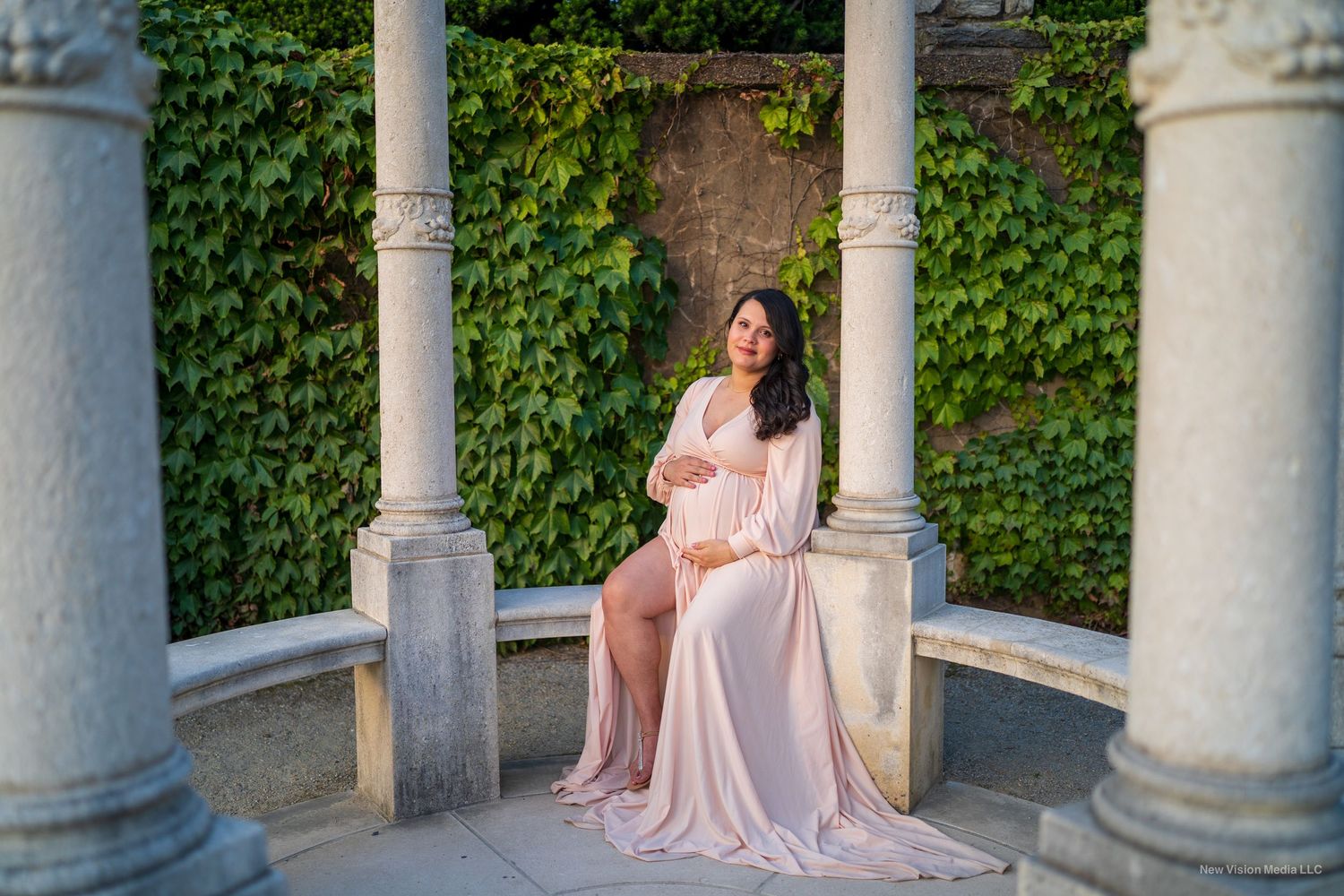 Maternity portrait in a pink flowing dress seated between classical stone columns with ivy-covered walls.