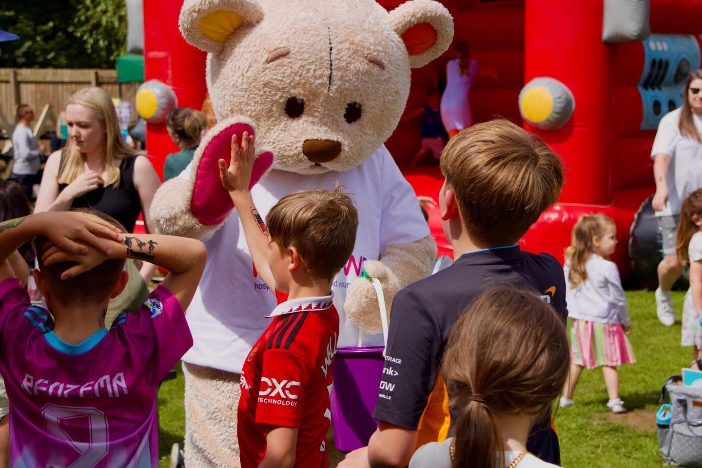 Children interact with a giant teddy bear mascot at a fun outdoor summer fair event with a bouncy castle.