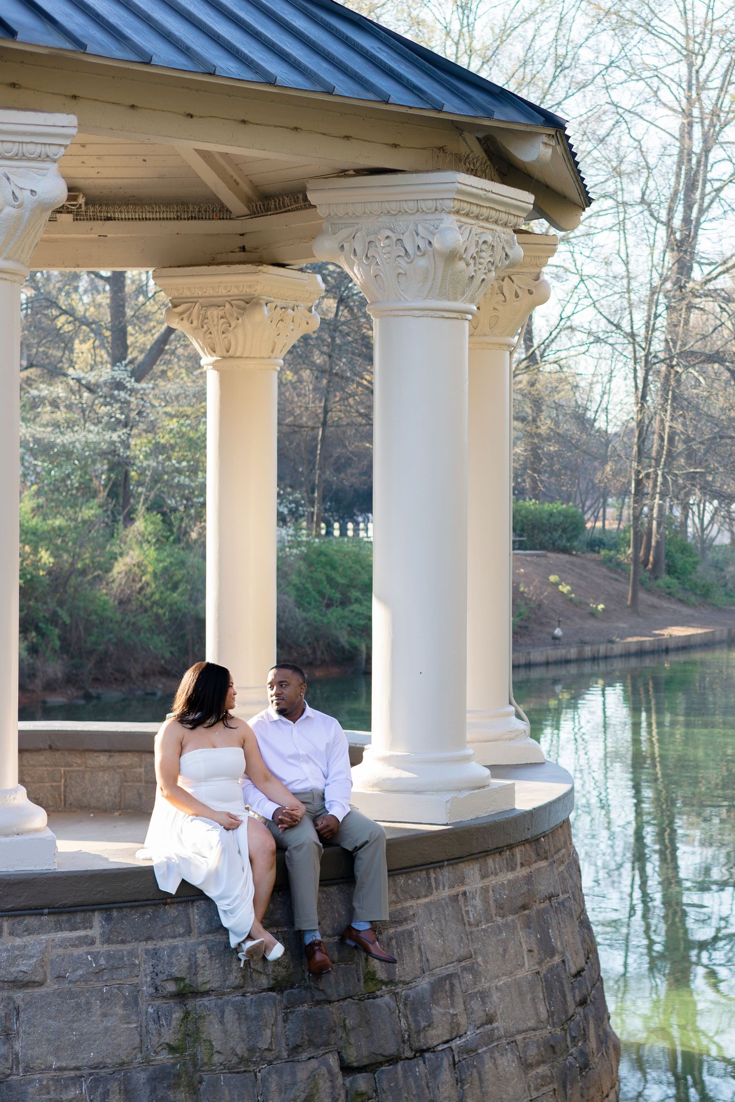 Couple sitting together under an elegant white gazebo by a serene pond, surrounded by trees in a romantic outdoor setting.