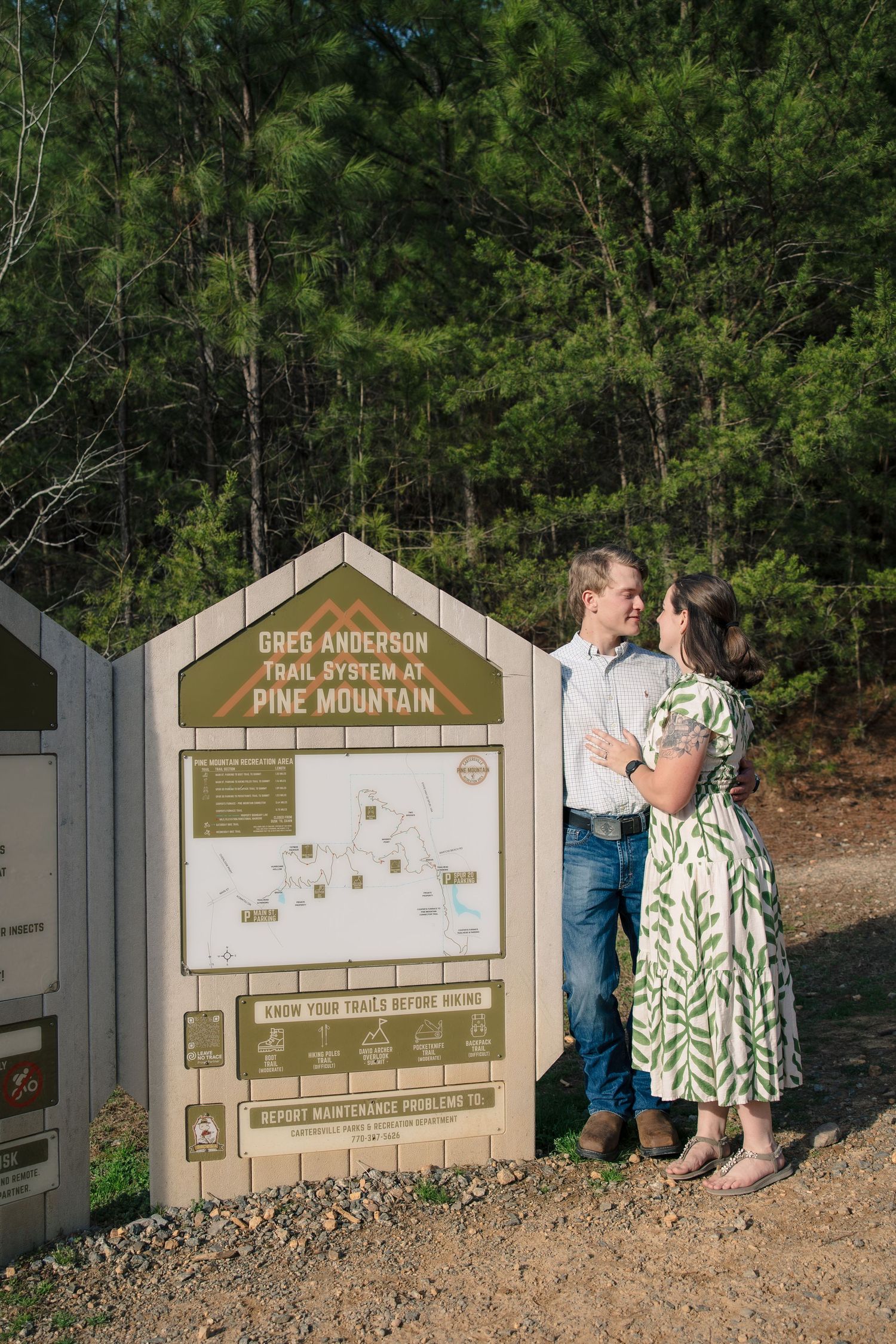 A couple stands by a Pine Mountain trail sign marker in a wooded area during a marriage proposal moment.