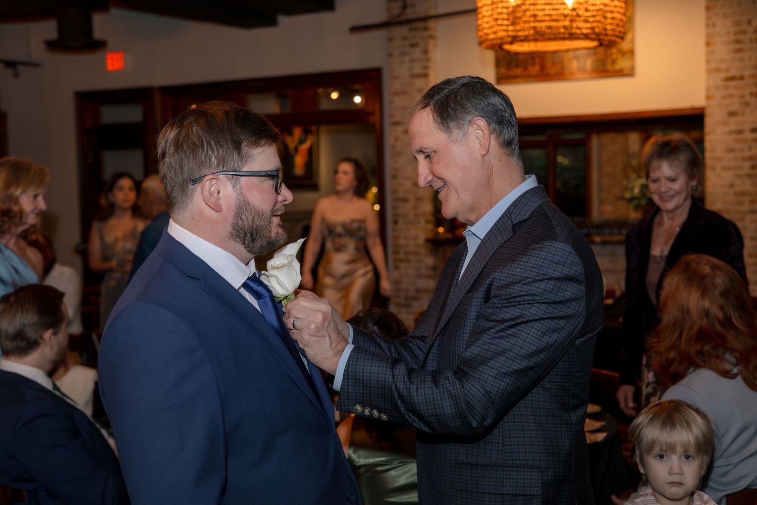 Emotional moment between two men in suits during wedding reception in brick-walled venue.