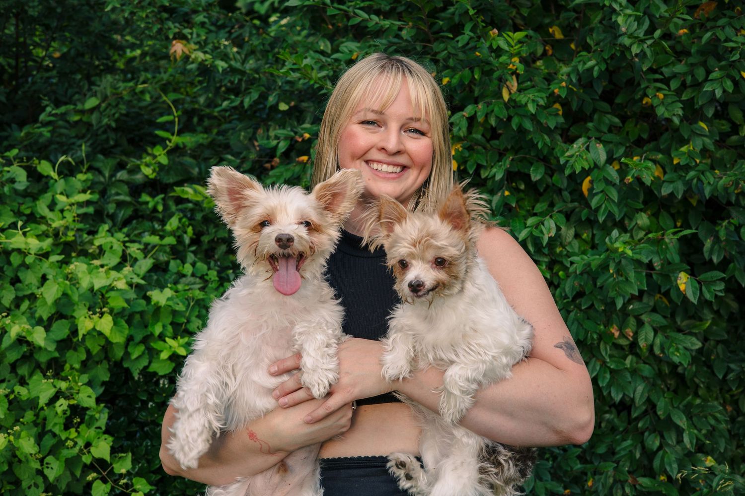 A person in a black top smiling while holding two small fluffy white dogs against green foliage.