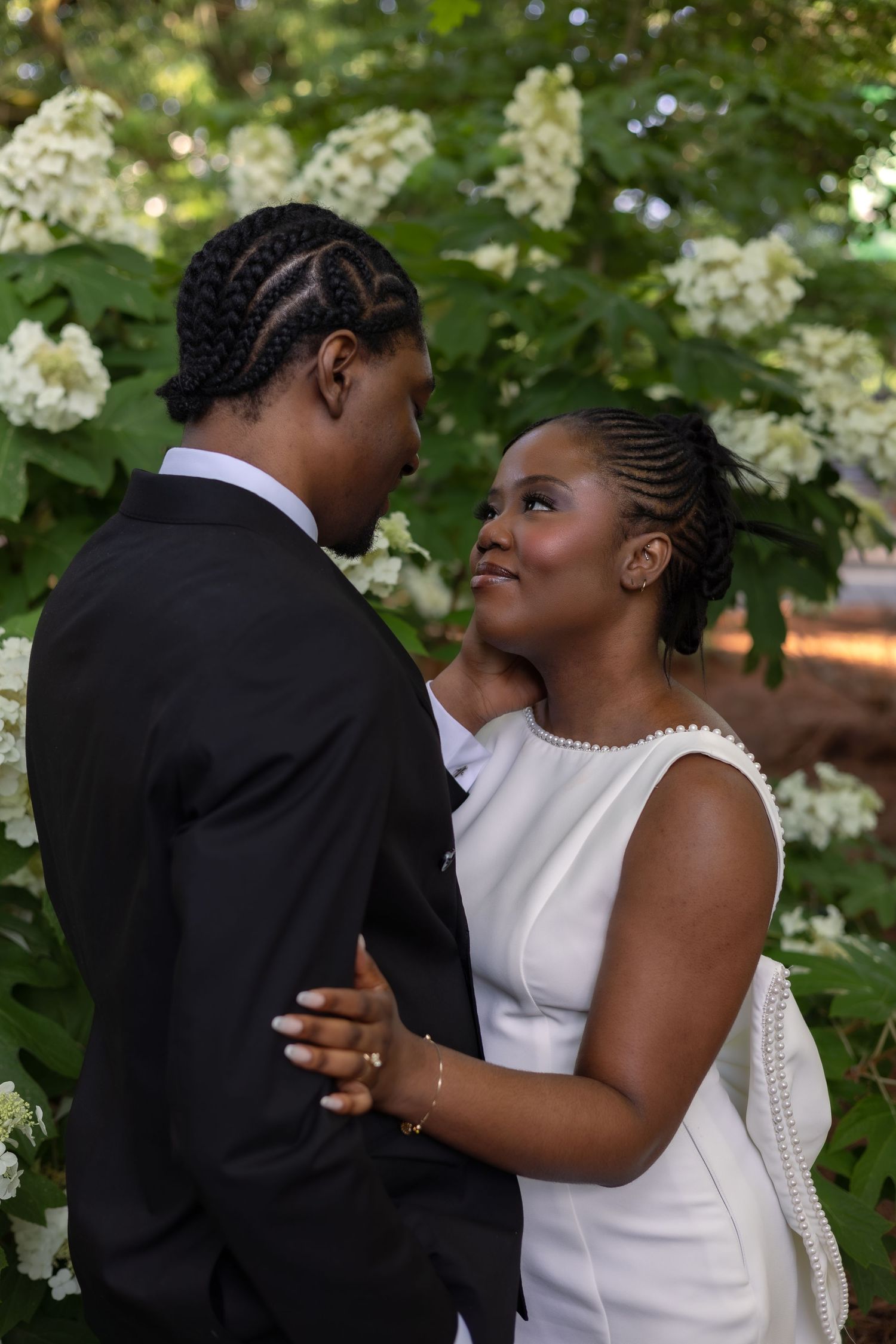 A Black couple in wedding attire embraces outdoors among white hydrangeas, the bride gazing lovingly at the groom.