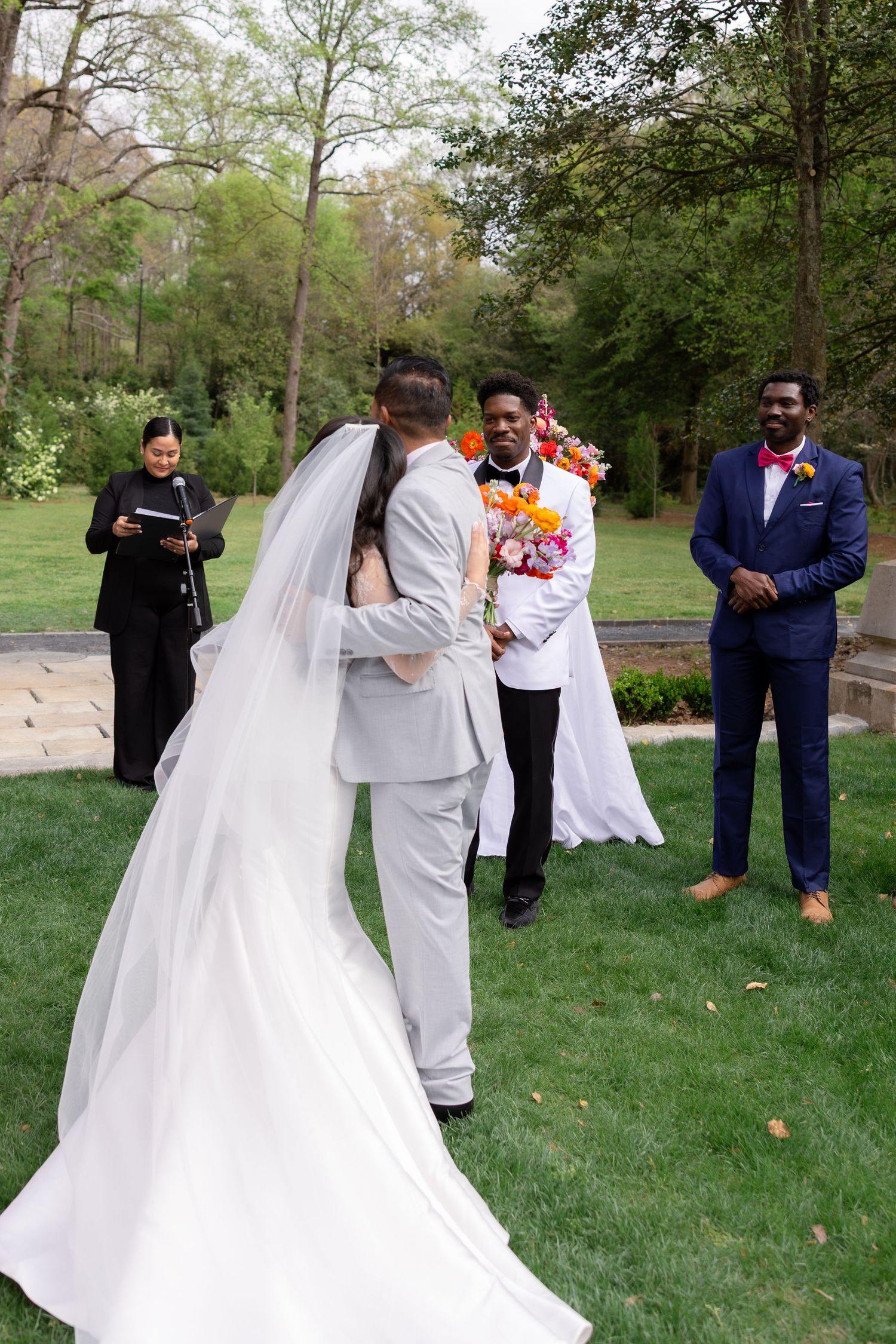 Outdoor wedding ceremony on green lawn with bride in white gown, groom in gray suit, officiant, and groomsmen.