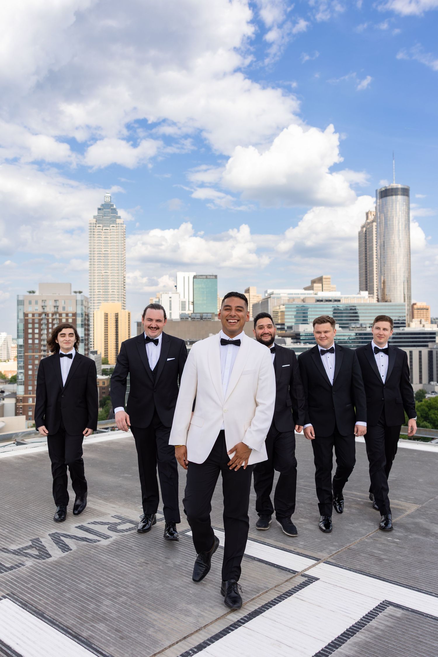 Groom in white tuxedo jacket leads groomsmen in black tuxedos on rooftop with Atlanta city skyline.