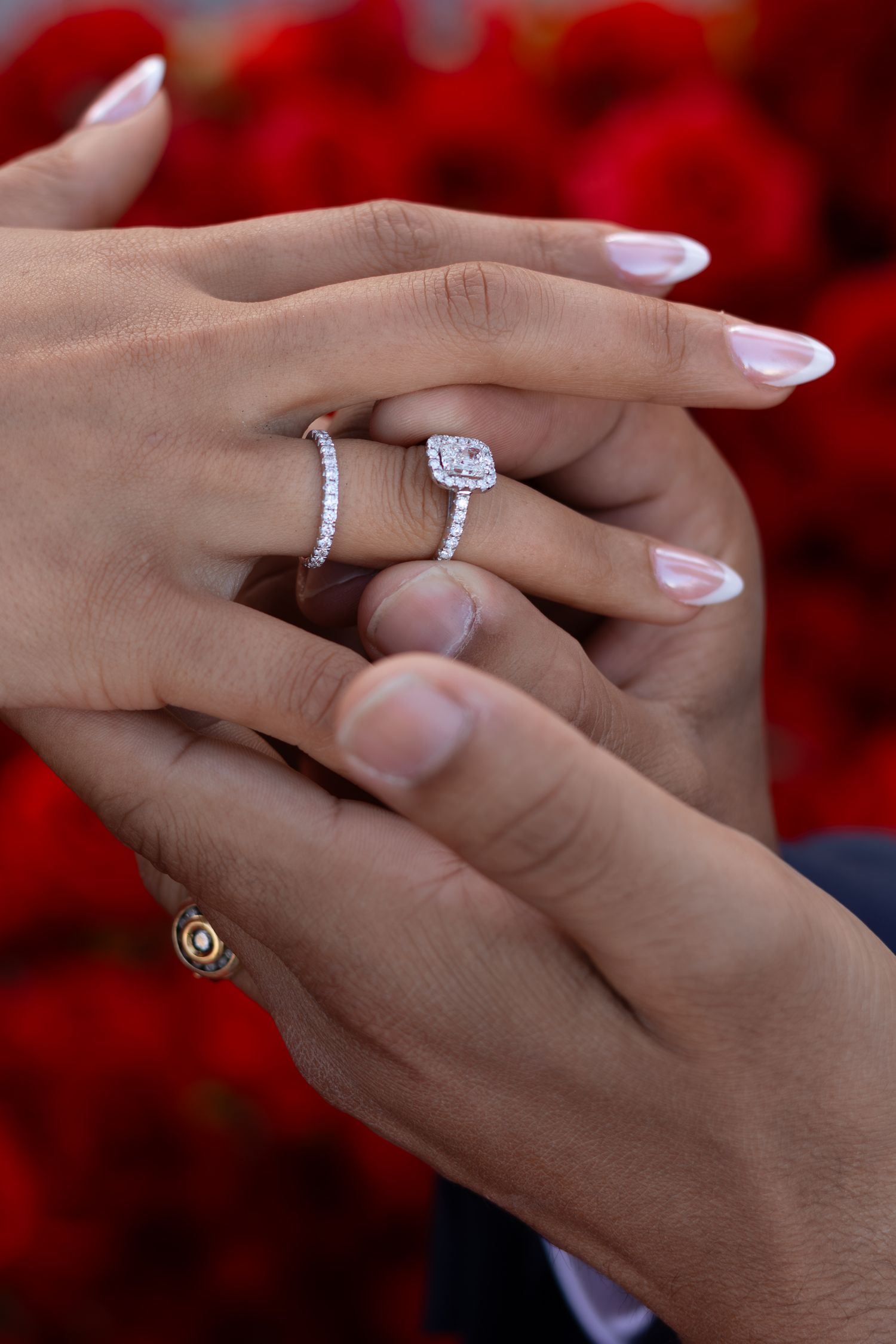 Close-up of hands with a diamond halo engagement ring and wedding band, with red roses in the background.