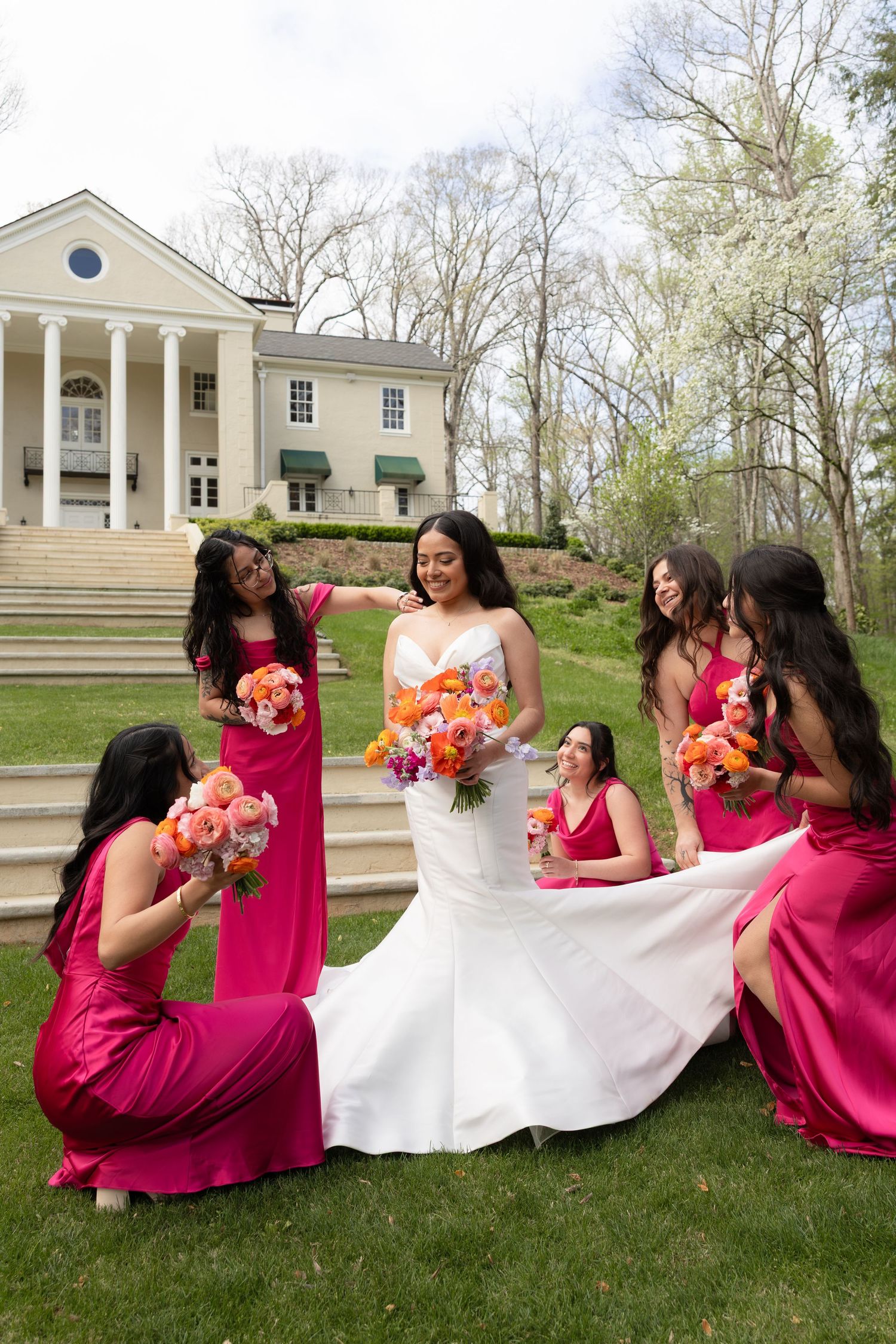 Bride in white gown surrounded by bridesmaids in pink dresses holding orange bouquets outside elegant venue.