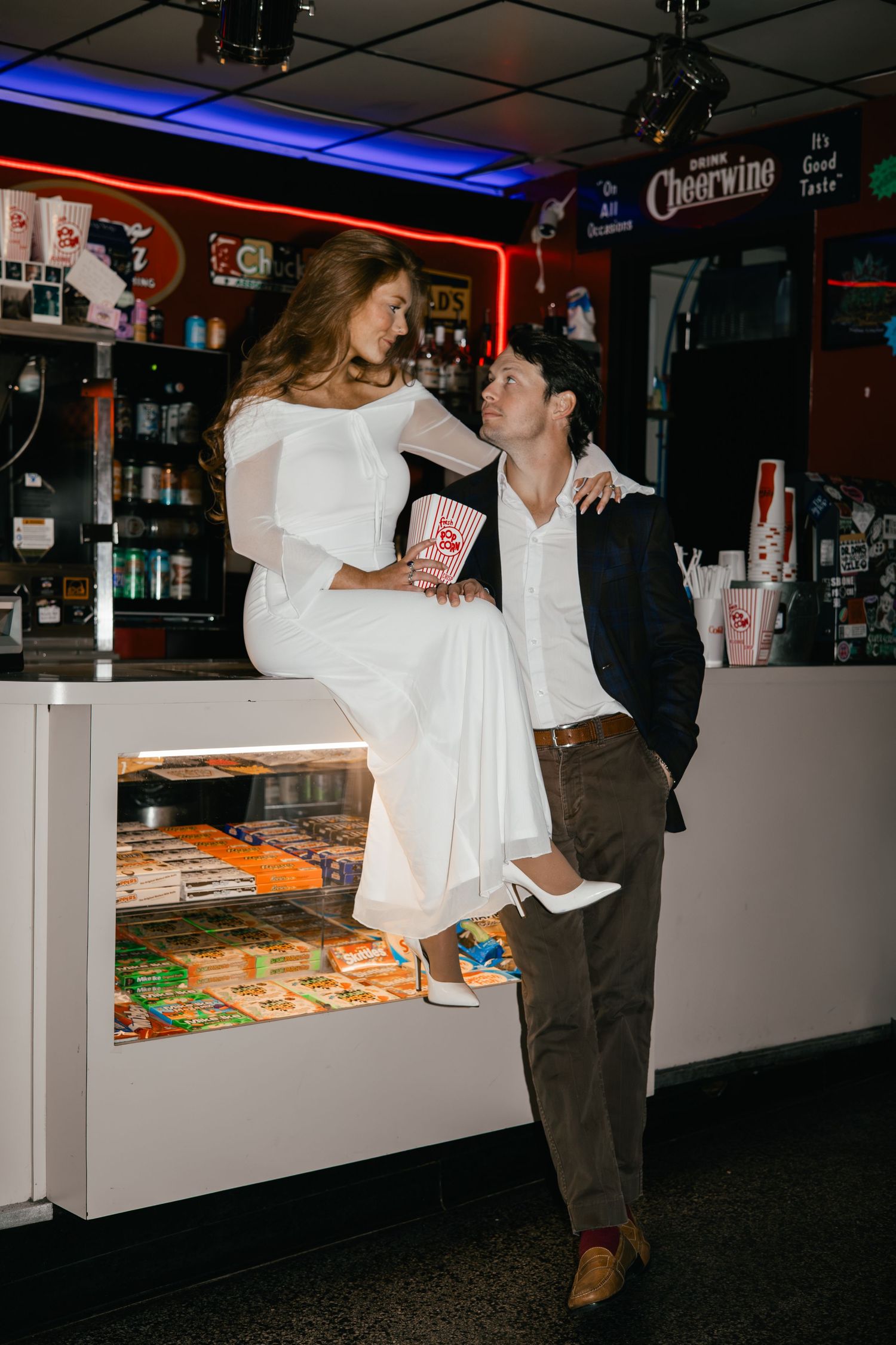 Two people, a woman in a white dress and a man in casual clothes, are embracing in what appears to be a bar or restaurant setting.