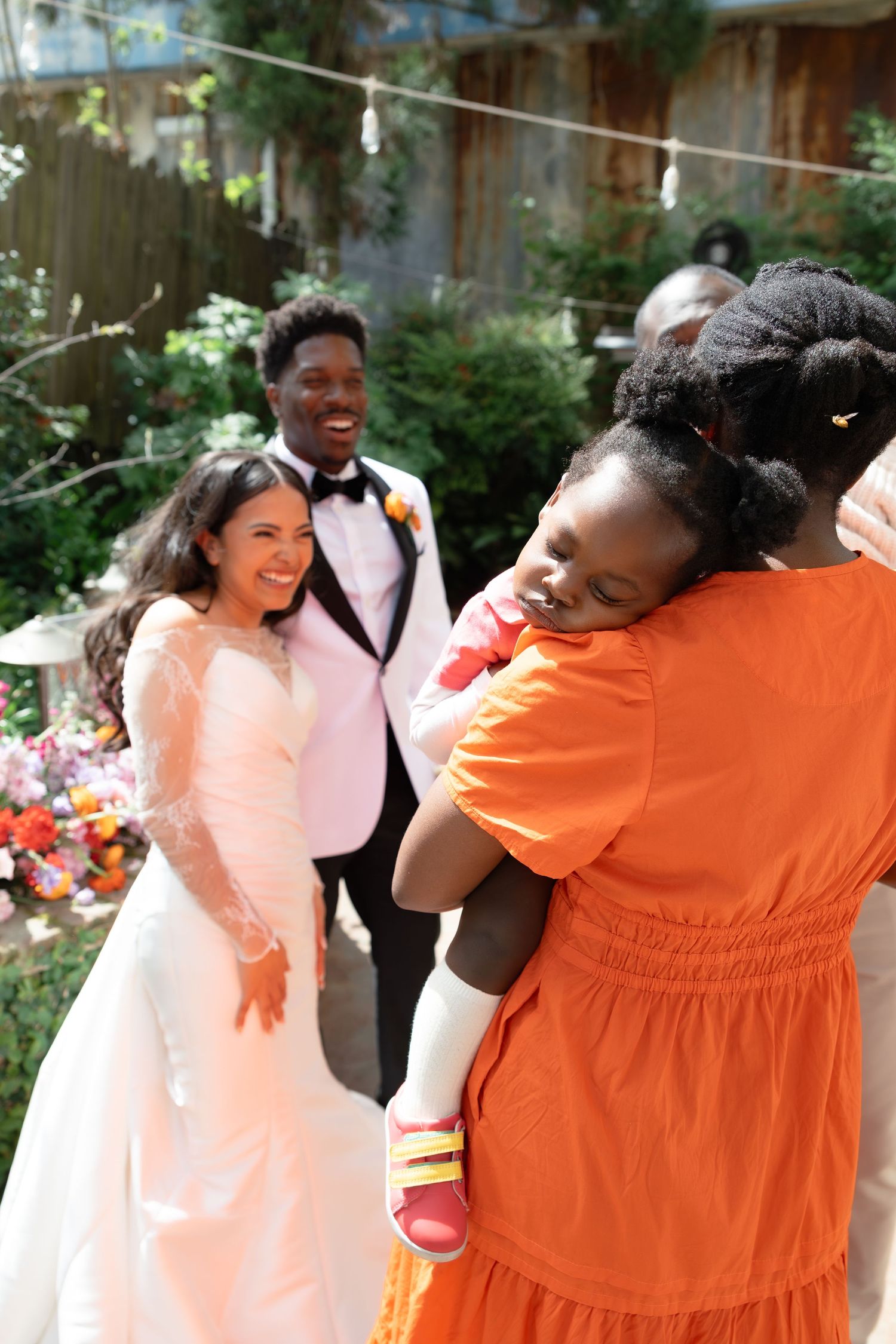 Bride and groom smile at guests during outdoor wedding reception, while woman in orange dress holds a young child.