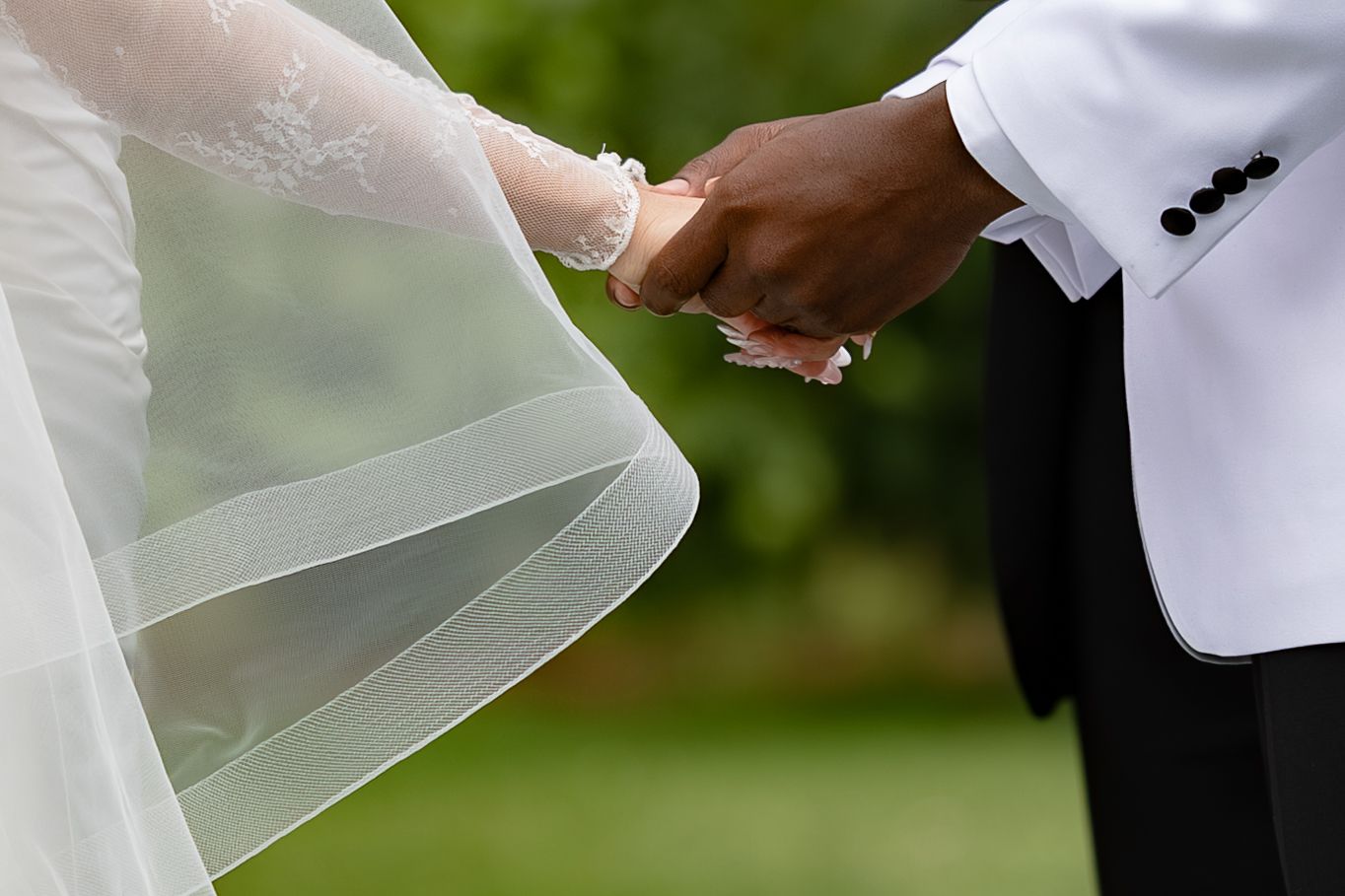 Interracial couple holding hands at wedding, bride in white gown with veil, groom in white tuxedo jacket.