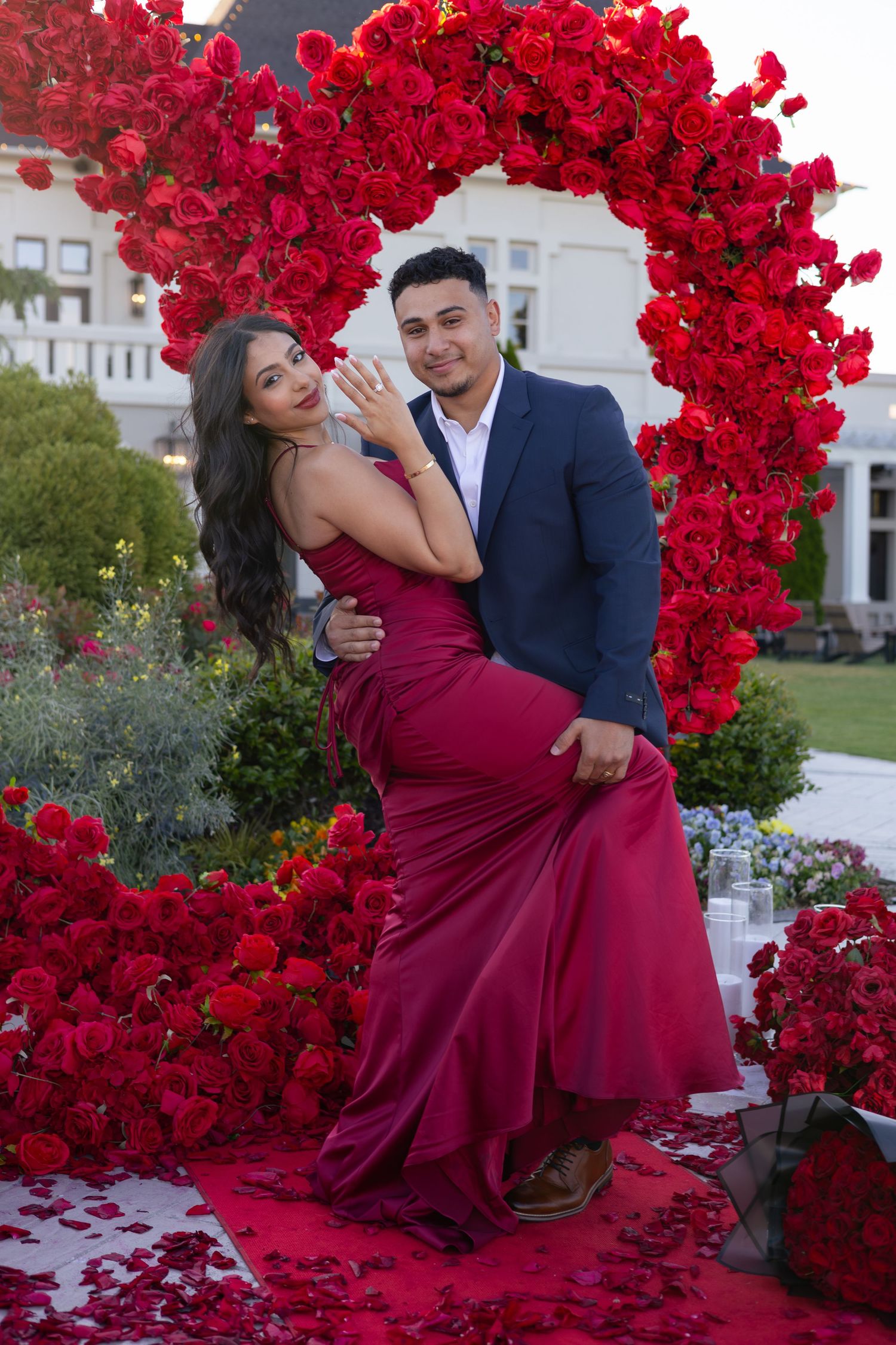 Couple posing in front of red rose heart arch during romantic outdoor proposal, woman showing engagement ring.
