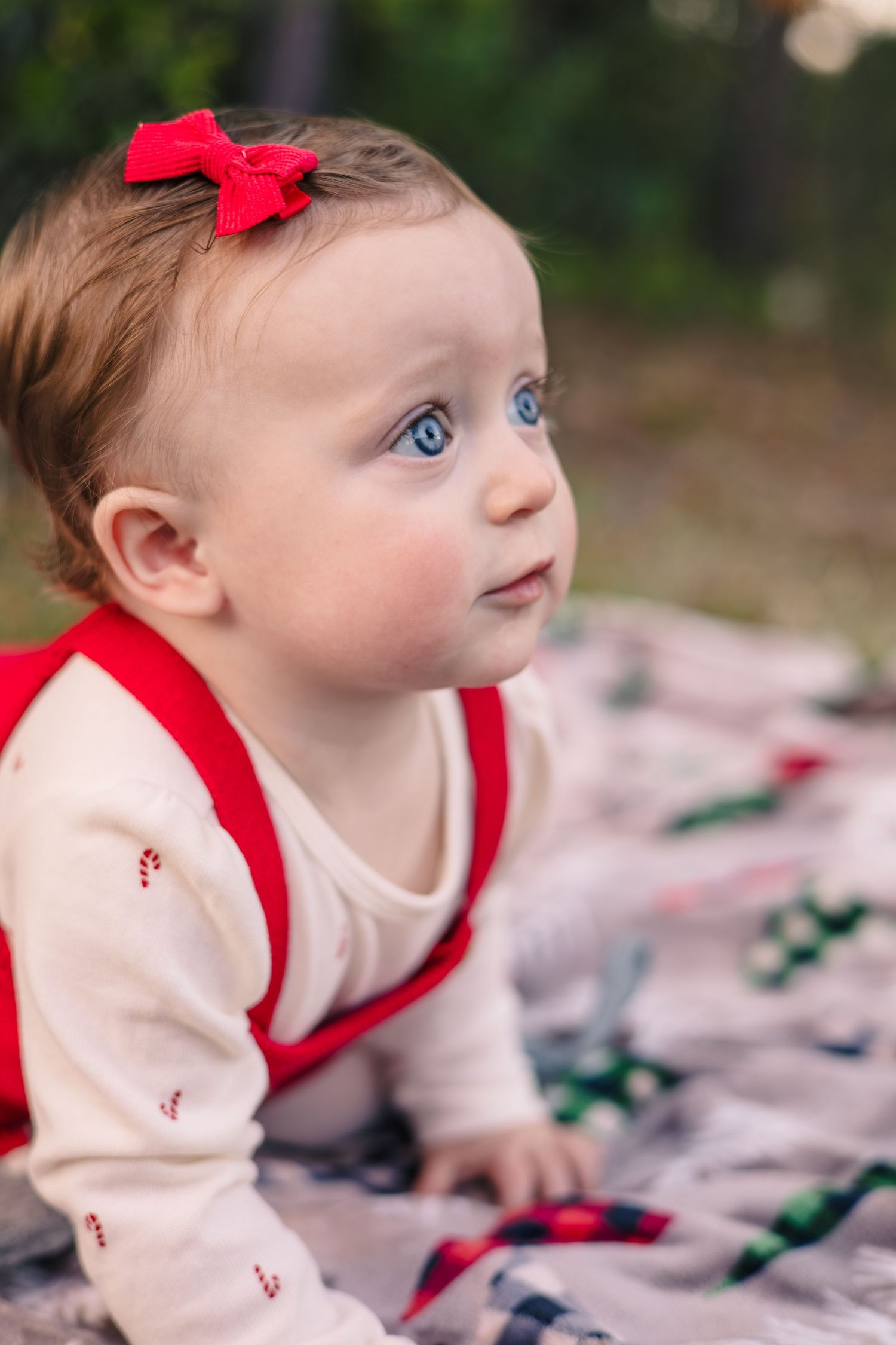 Baby wearing white and red outfit with bow lying on blanket during outdoor photo session.