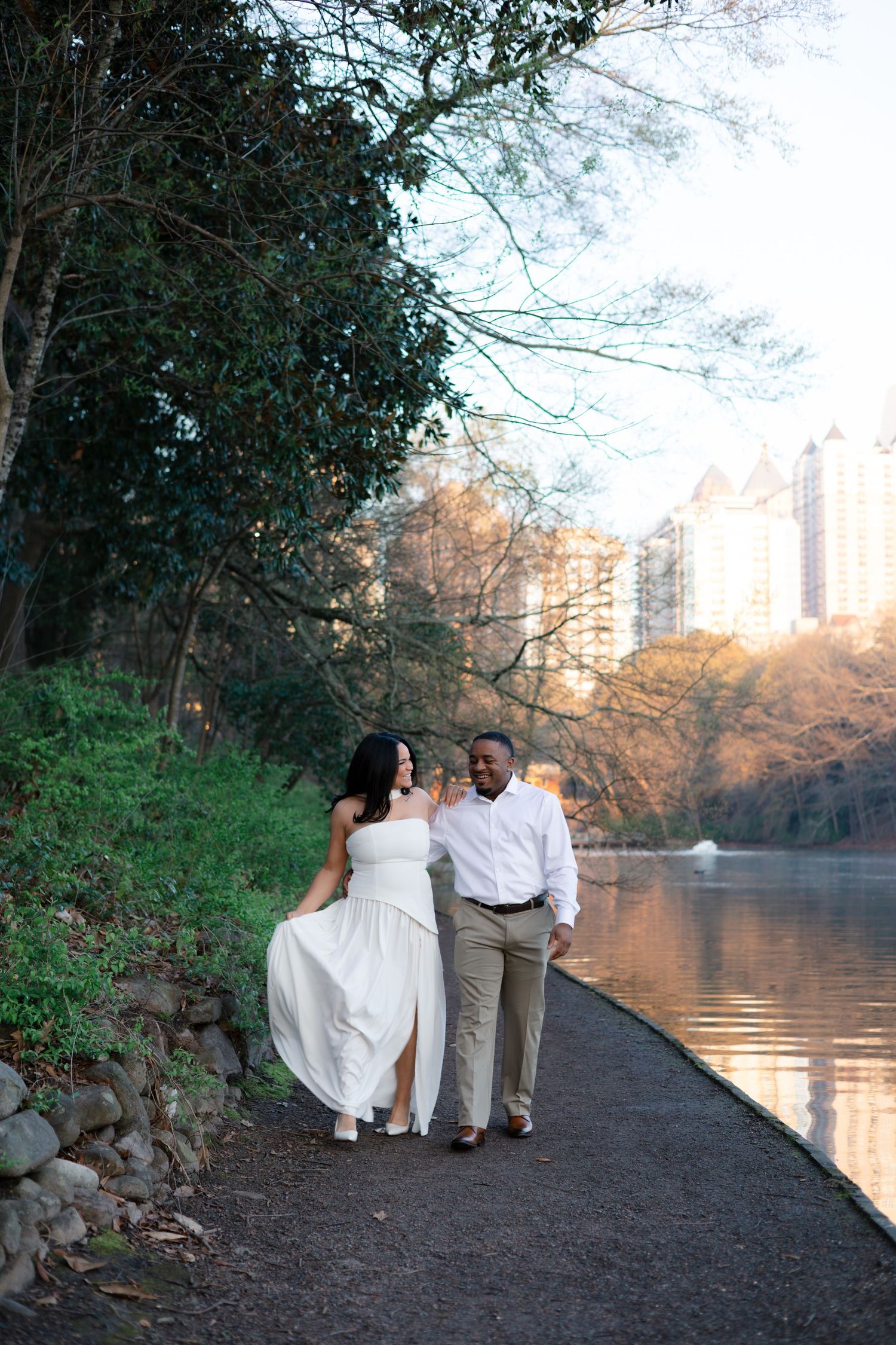 Three figures walk along a scenic path beside a tranquil lake with city buildings visible in the autumn landscape.