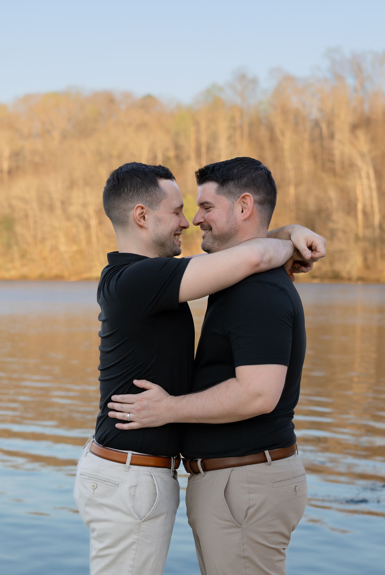 Two people in black shirts and khaki pants embrace lovingly by a serene lake during golden hour.