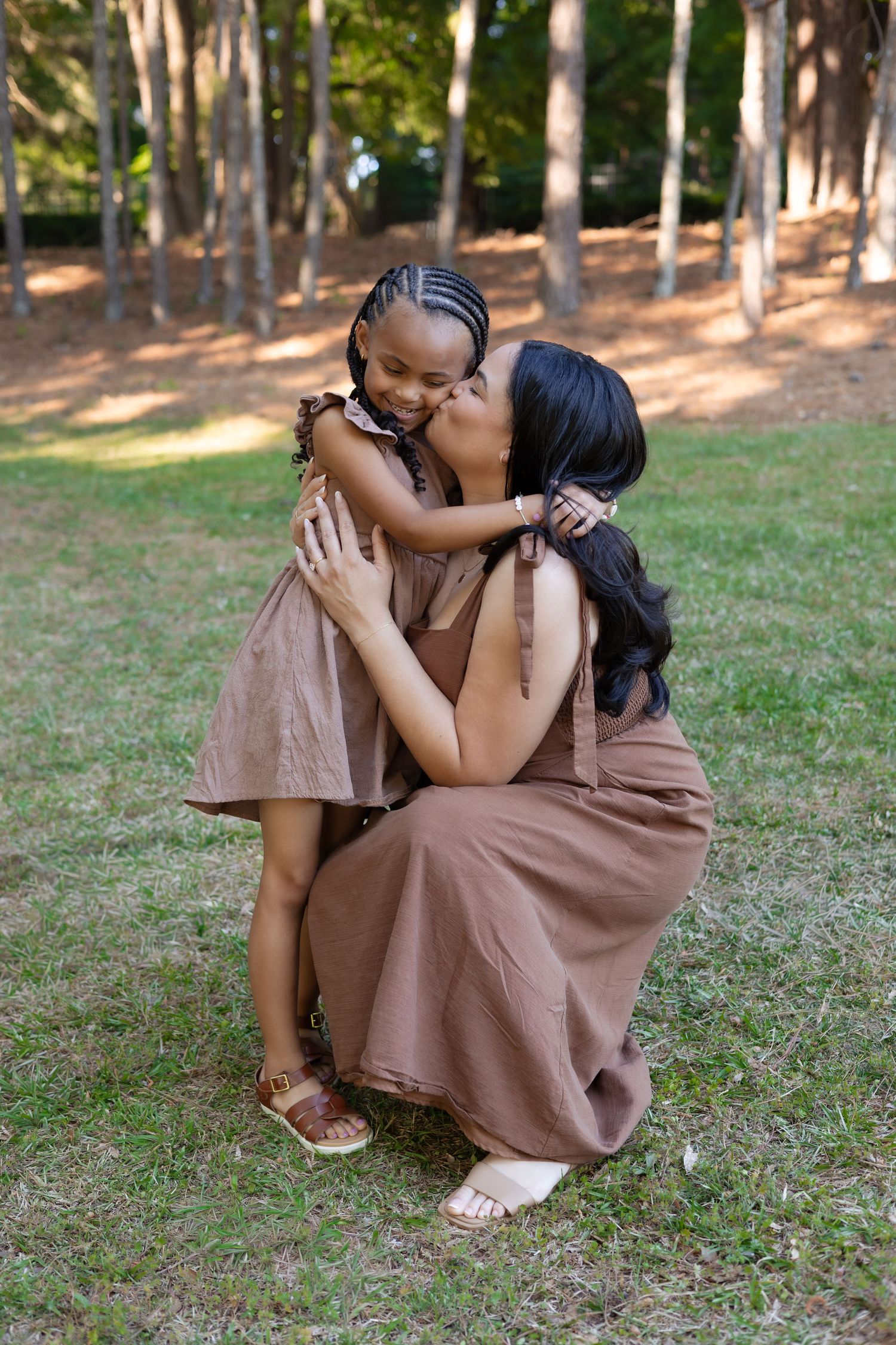 A mother crouches to kiss her young daughter in a park, both wearing matching brown dresses among tall trees.
